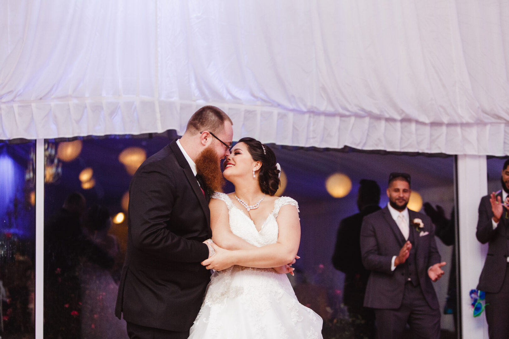 At a Brandywine Manor House Wedding, a couple in elegant wedding attire shares an enchanting moment on the dance floor, their joy illuminated by the warm applause of delighted guests in the background.