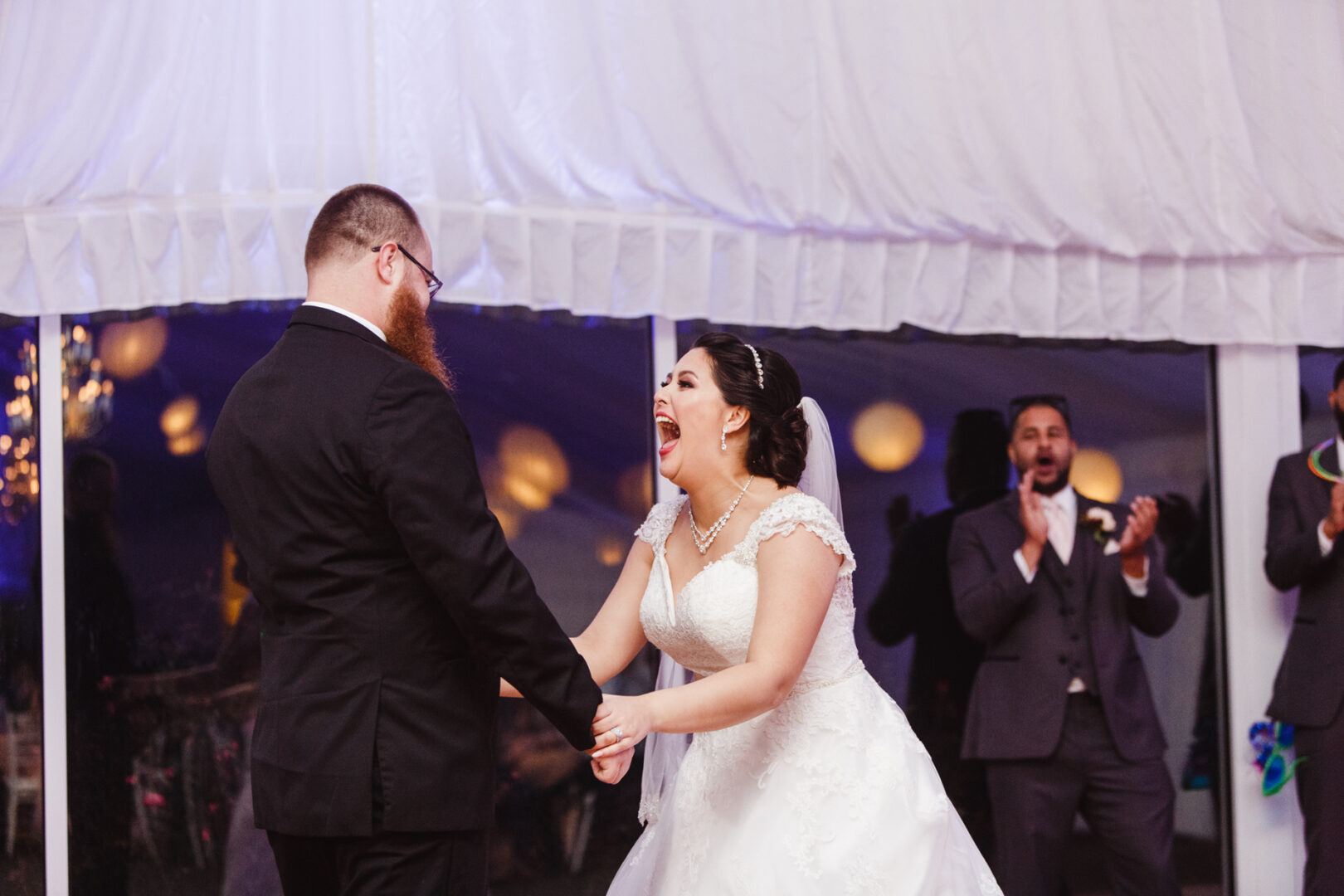 At a Brandywine Manor House wedding, a bride and groom hold hands and smile joyfully under a white tent, with applauding guests in the background.