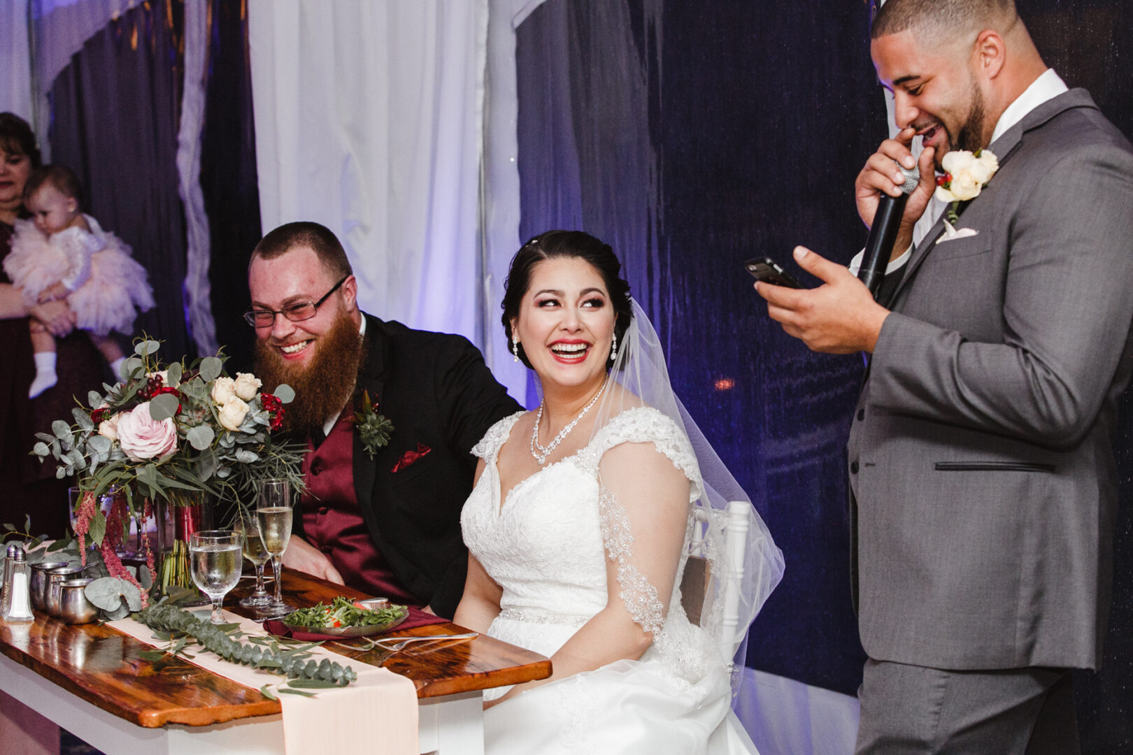 At a charming Brandywine Manor House wedding, a bride and groom sit smiling at a table with stunning floral arrangements. Beside them, a man in a suit enthusiastically speaks into a microphone while holding his phone, capturing the joyful moment.