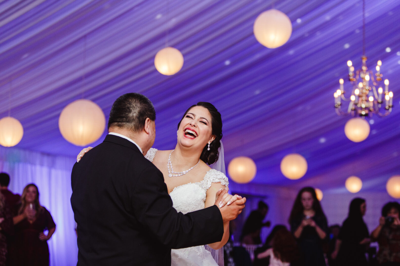 At a charming Brandywine Manor House wedding, a couple dances joyfully under a canopy adorned with twinkling lights and an elegant chandelier, while guests watch in delight from the background.