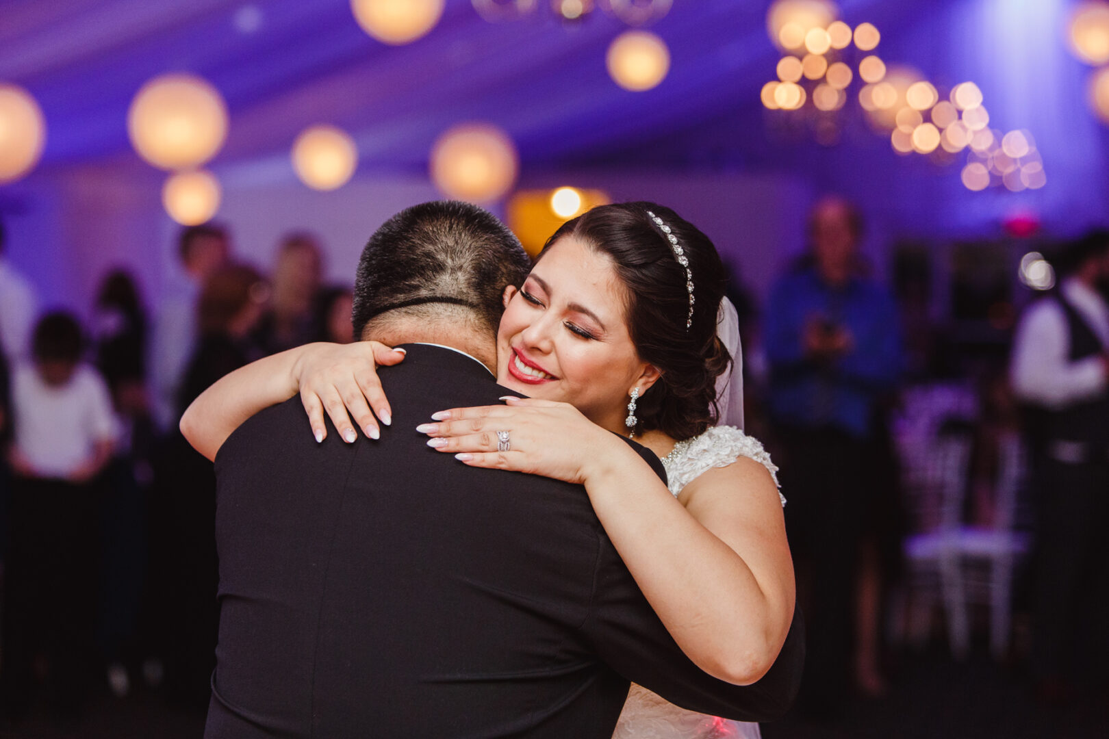 At a Brandywine Manor House wedding, a bride in a white dress embraces a man in a black suit while they dance. The backdrop is beautifully adorned with string lights and spherical lanterns, enhancing the enchanting atmosphere.