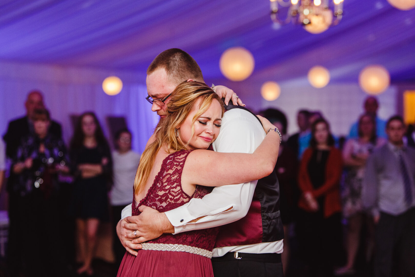 At a Brandywine Manor House wedding, a woman in a maroon dress and a man in formal attire embrace closely on the dance floor. Guests watch them under soft, white lighting with hanging round decorations, adding to the enchanting atmosphere.