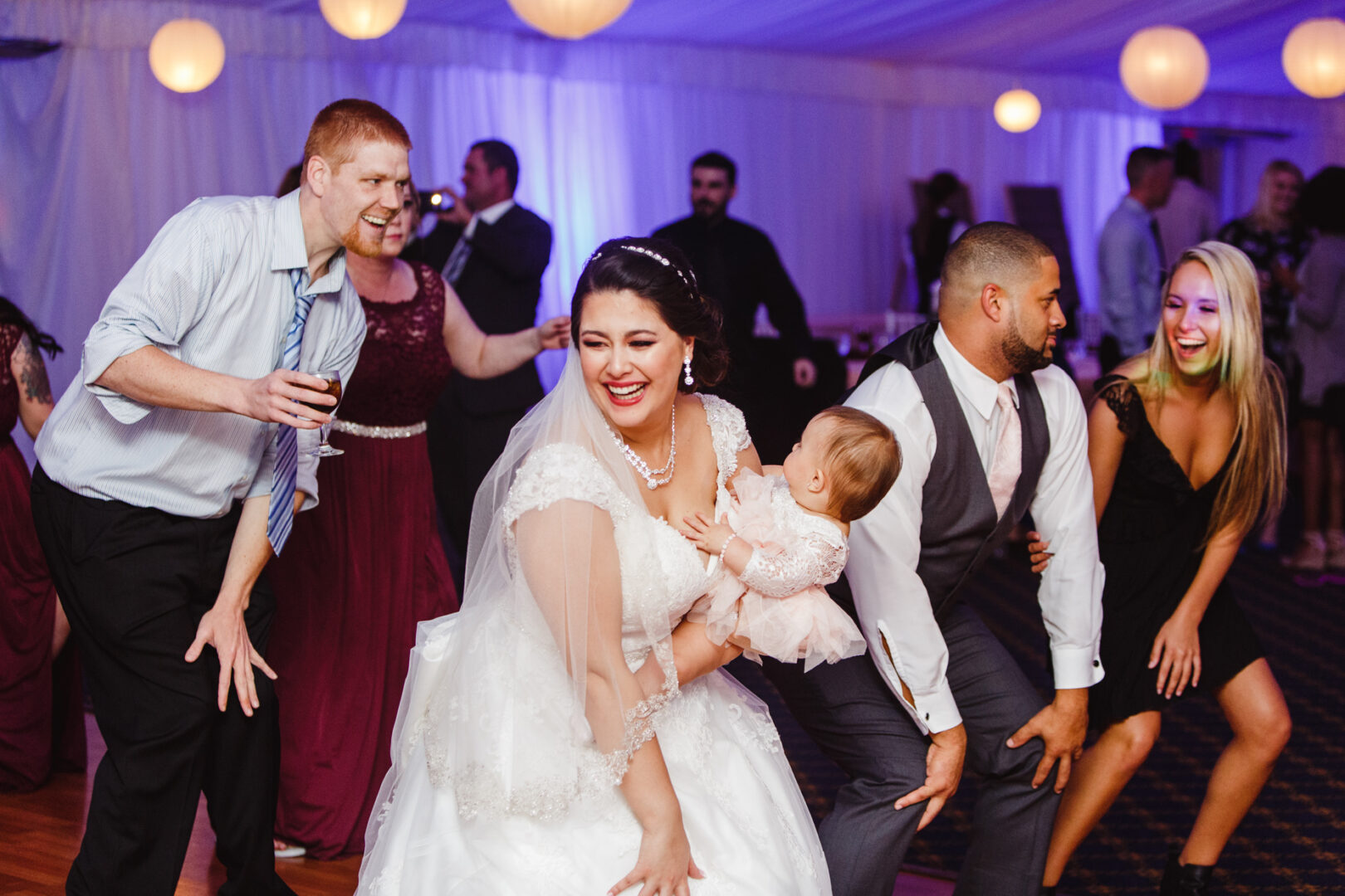 At a Brandywine Manor House wedding, the bride and groom dance energetically while holding a baby, surrounded by smiling guests under a tent with hanging lights.