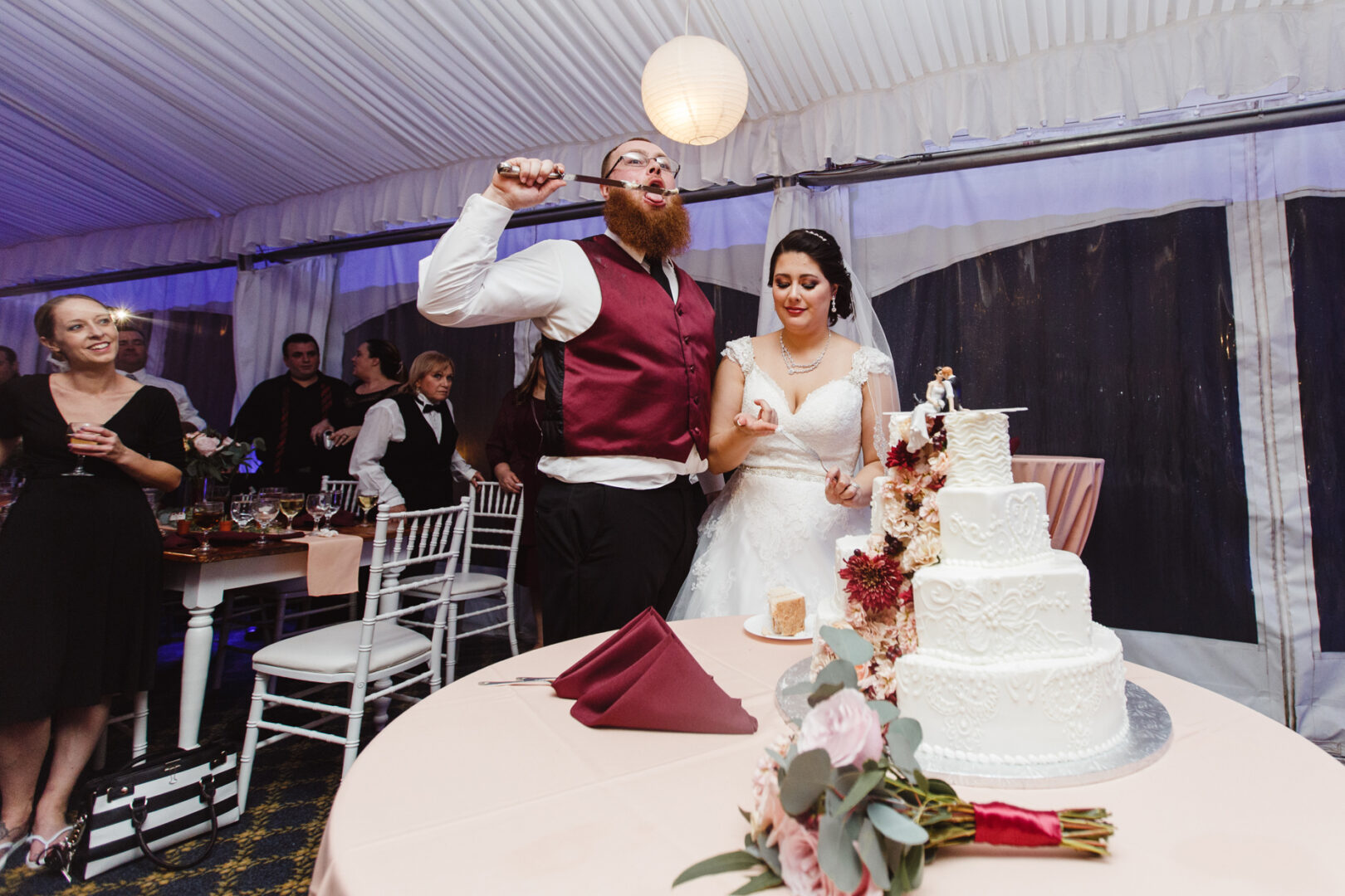 At the Brandywine Manor House Wedding, a groom holding a microphone stands beside his bride in front of a tiered cake. Surrounded by guests, they celebrate in the beautifully decorated reception area.