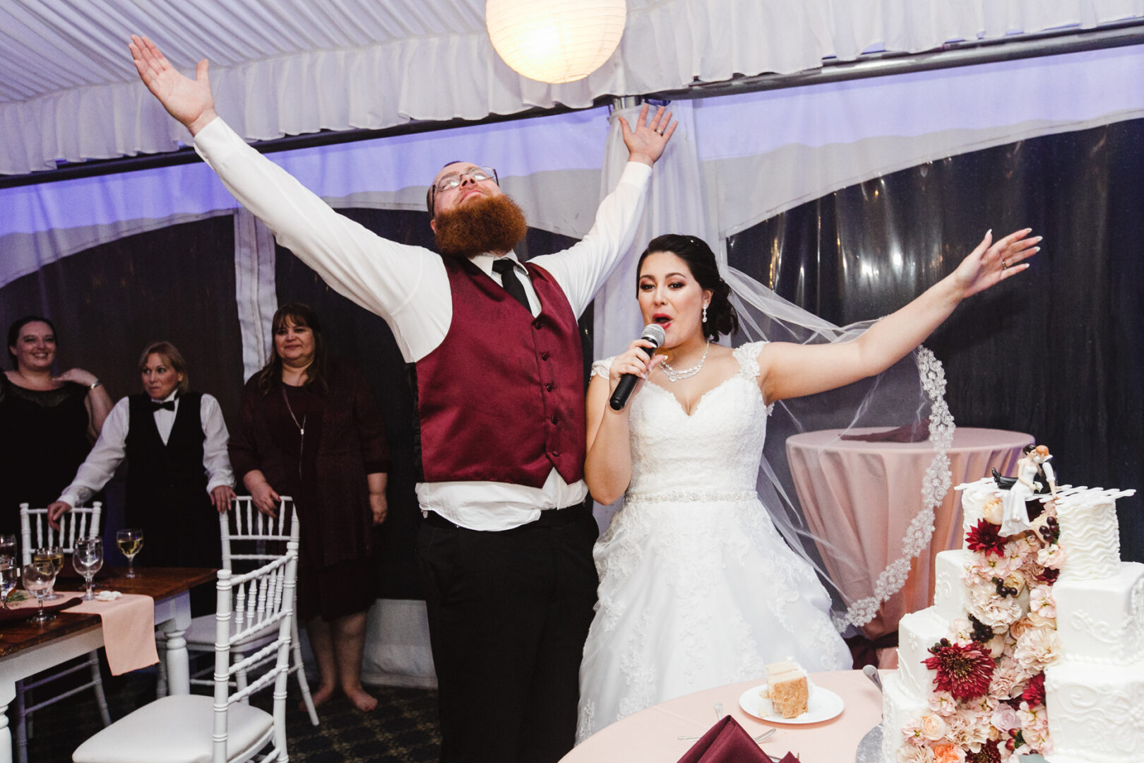 At a Brandywine Manor House Wedding, the bride in a stunning wedding dress holds a microphone as the groom in a vest joyously raises his arms. In the foreground, a beautifully crafted wedding cake stands, with guests seated and enjoying the celebration in the background.