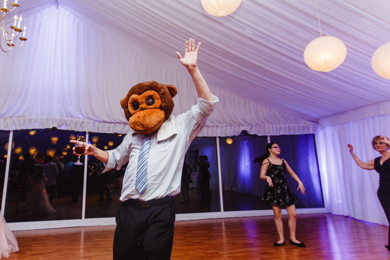 At a Brandywine Manor House wedding, a person in a monkey mask dances wildly under the glow of hanging lamps, drink in hand. Nearby, two others join the fun inside the lively event tent.