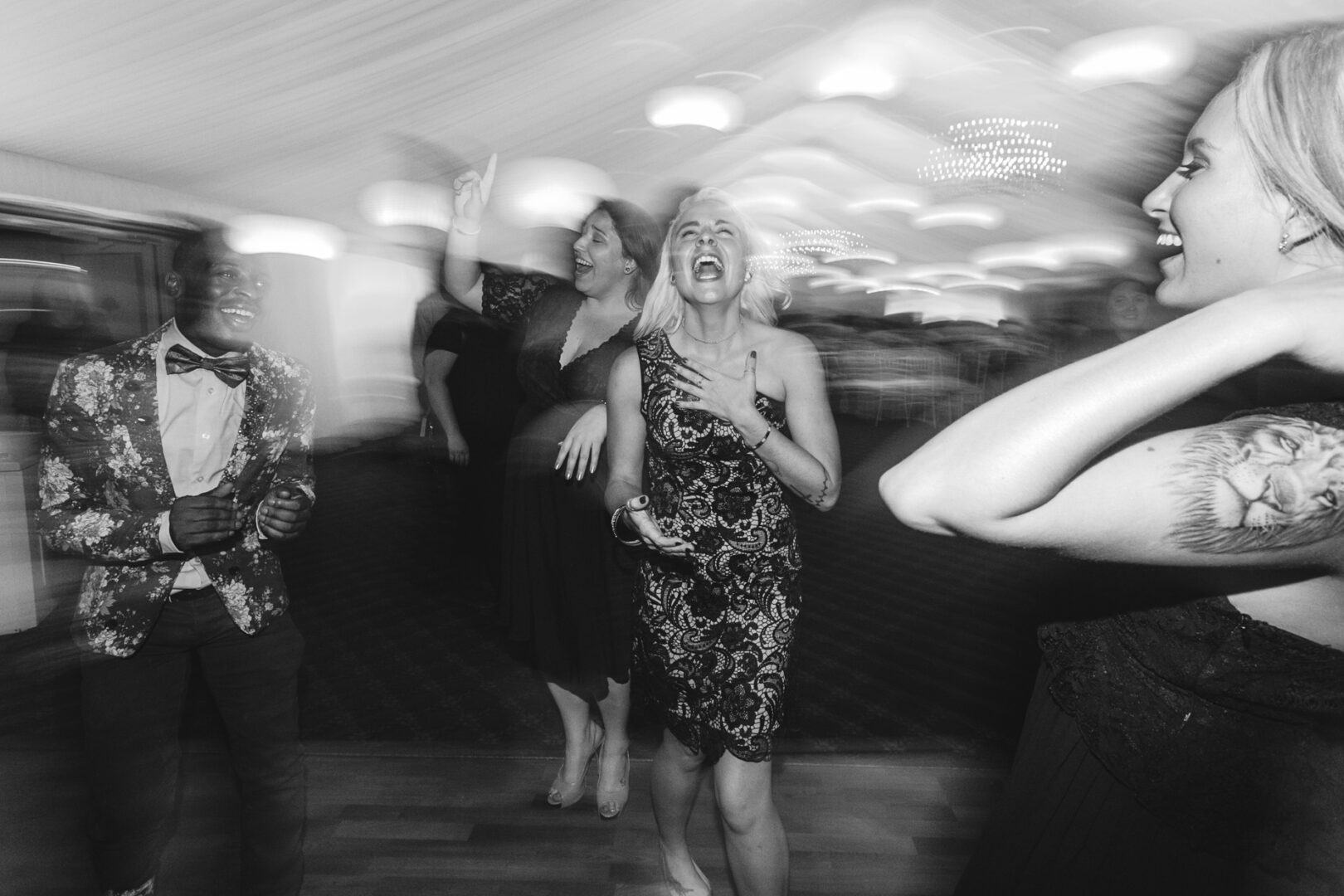 At Brandywine Manor House, a group of people in formal attire joyfully dance in a blurred black-and-white indoor setting, capturing the timeless essence of a wedding celebration.