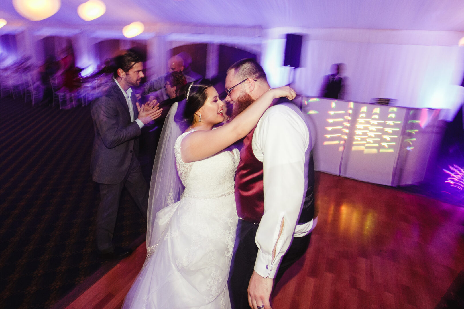 At the Brandywine Manor House wedding, a bride and groom share an intimate dance as a few guests join them on the softly lit floor, surrounded by a dreamy backdrop of purple and orange tones.