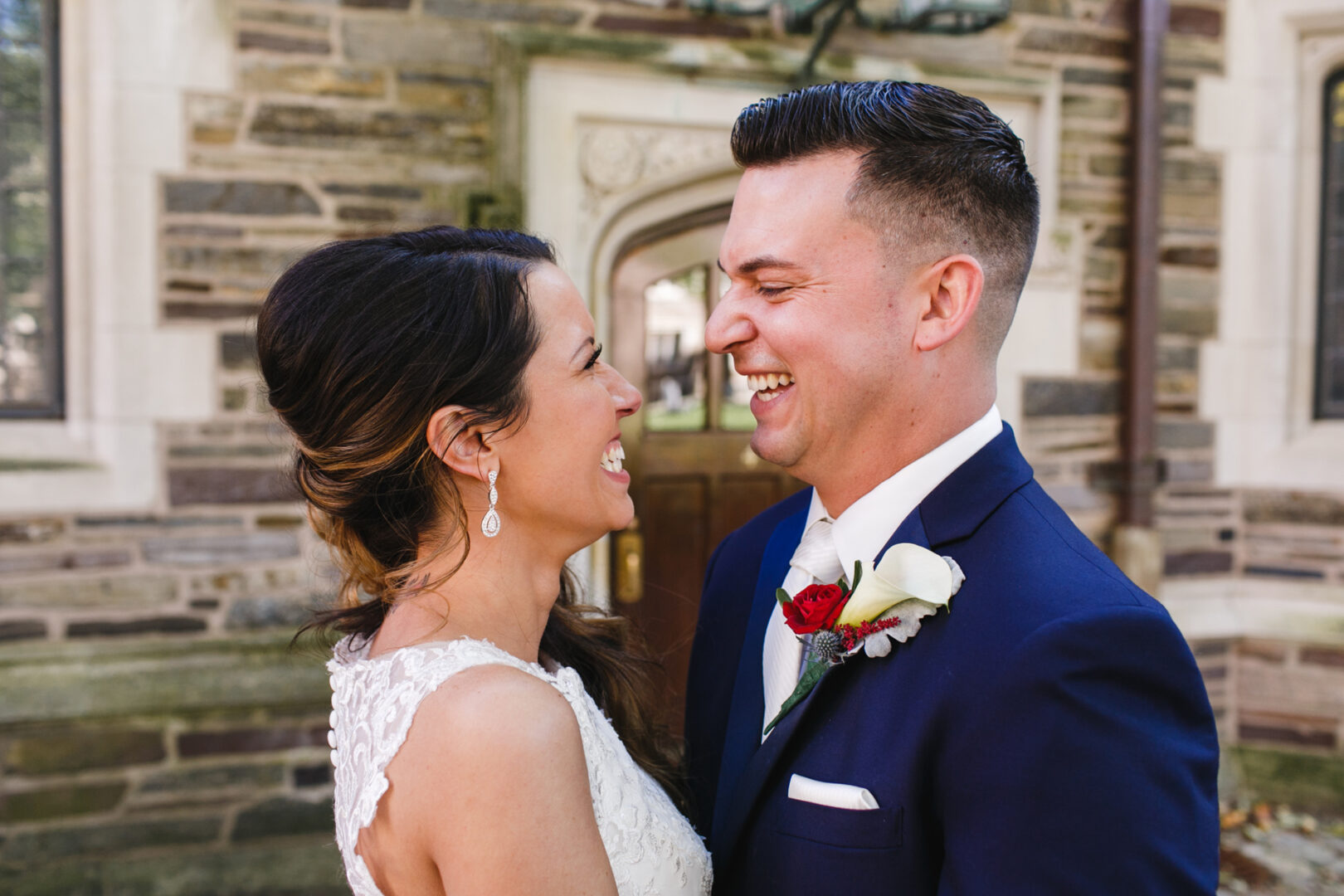 A bride and groom share a joyful moment outside a stone building, reminiscent of their elegant Windsor Ballroom wedding. The groom is dashing in a navy suit with a white boutonniere, while the bride stuns in her lace dress and long earrings.