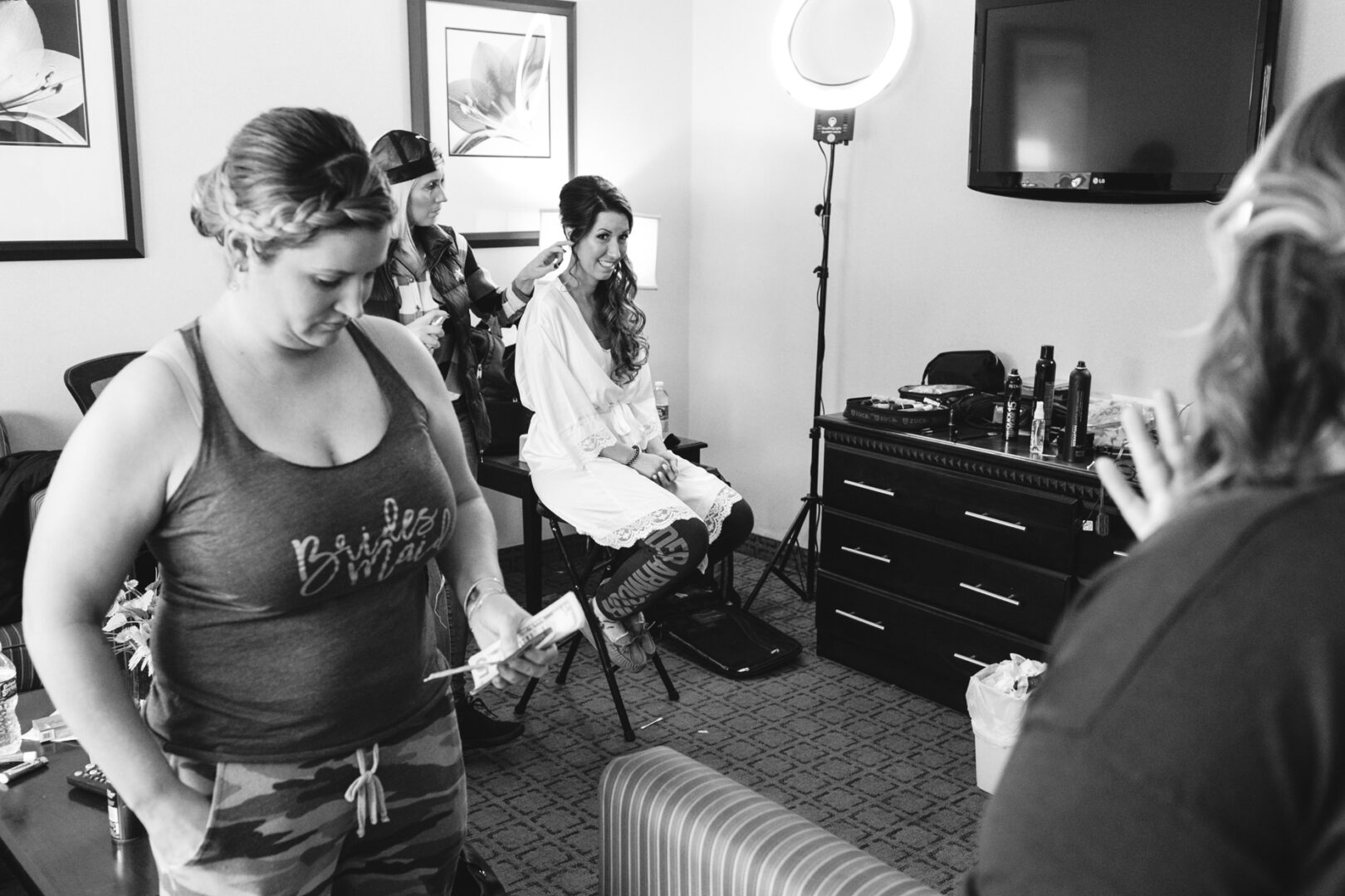 In a hotel room buzzing with excitement, a bride-to-be gets her hair styled for her Windsor Ballroom wedding. She's surrounded by friends, a ring light casting a glow over beauty products and the tell-tale "Bride's Maid" shirt that's ready for the festivities ahead.