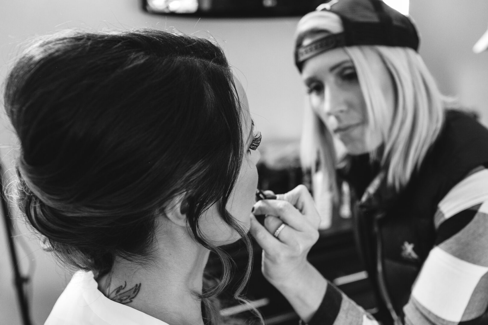 A woman with an updo hairstyle is having her makeup applied by another woman wearing a cap, preparing for what looks like a Windsor Ballroom wedding. The setting appears to be indoors, captured in timeless black and white.