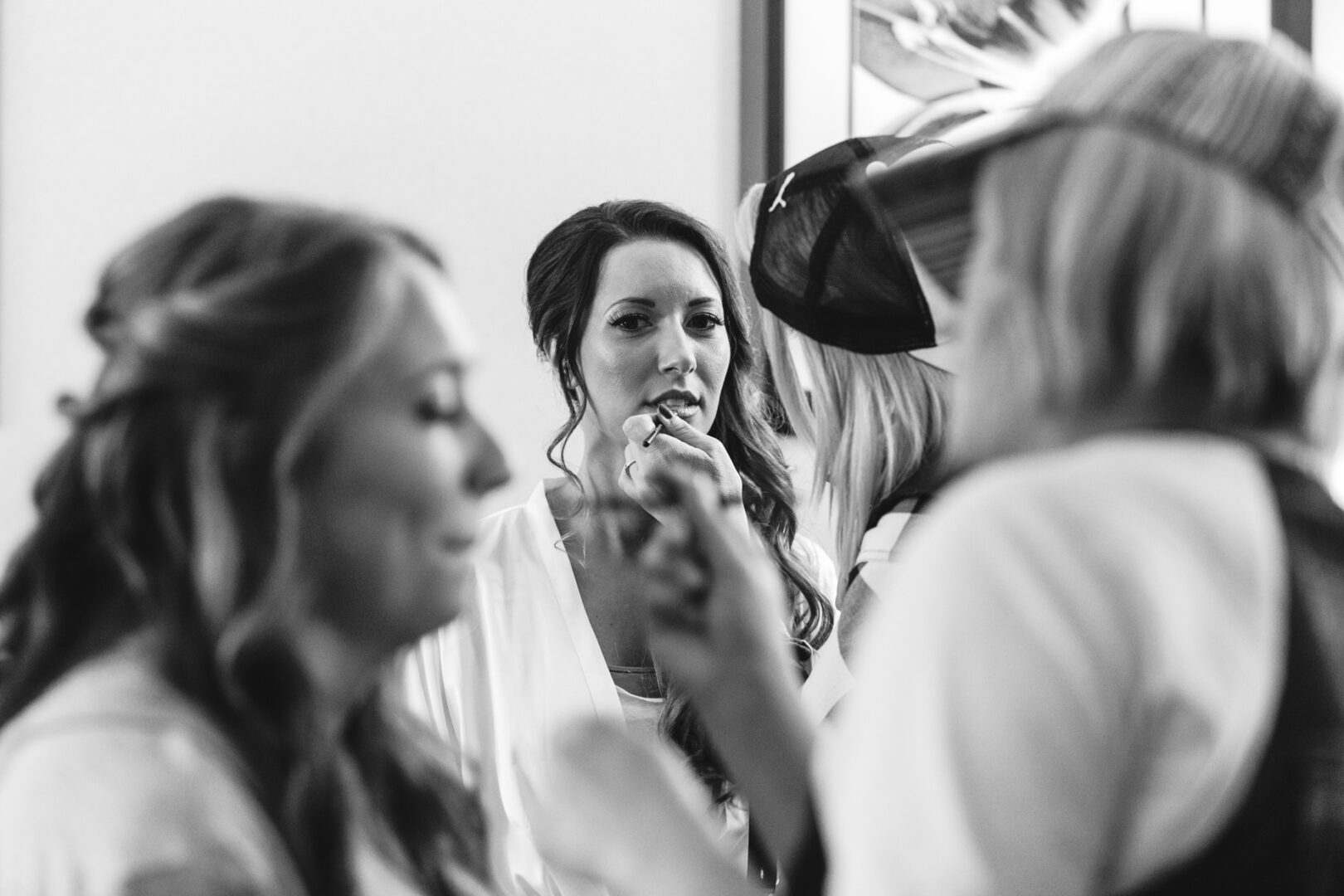 In the elegant Windsor Ballroom, a woman sits gracefully as her makeup is delicately applied, surrounded by an attentive gathering. The timeless black and white scene captures a moment filled with anticipation on this special wedding day.