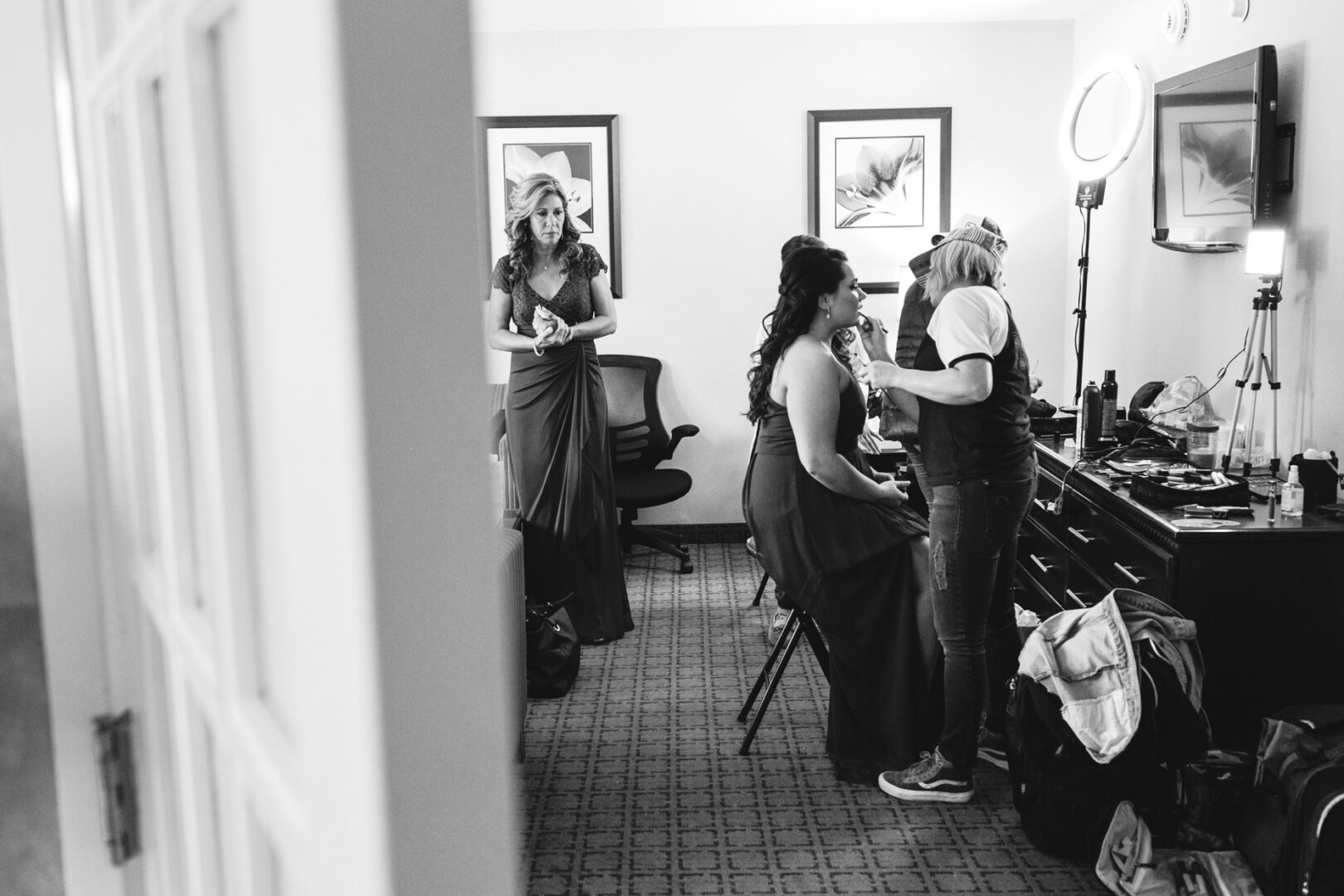 In a black and white photo, a woman sits serenely as another applies her makeup, preparing for the Windsor Ballroom Wedding. In the background, a woman in a dress stands gracefully. The room features two framed pictures, an open door, and a well-lit makeup station.