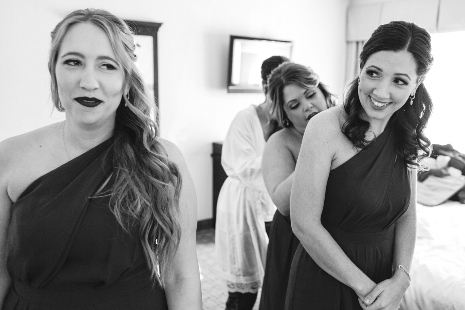 Three women in elegant dresses prepare in a room, with one adjusting another's outfit. They seem poised for the Windsor Ballroom wedding, captured beautifully in a timeless black and white image.