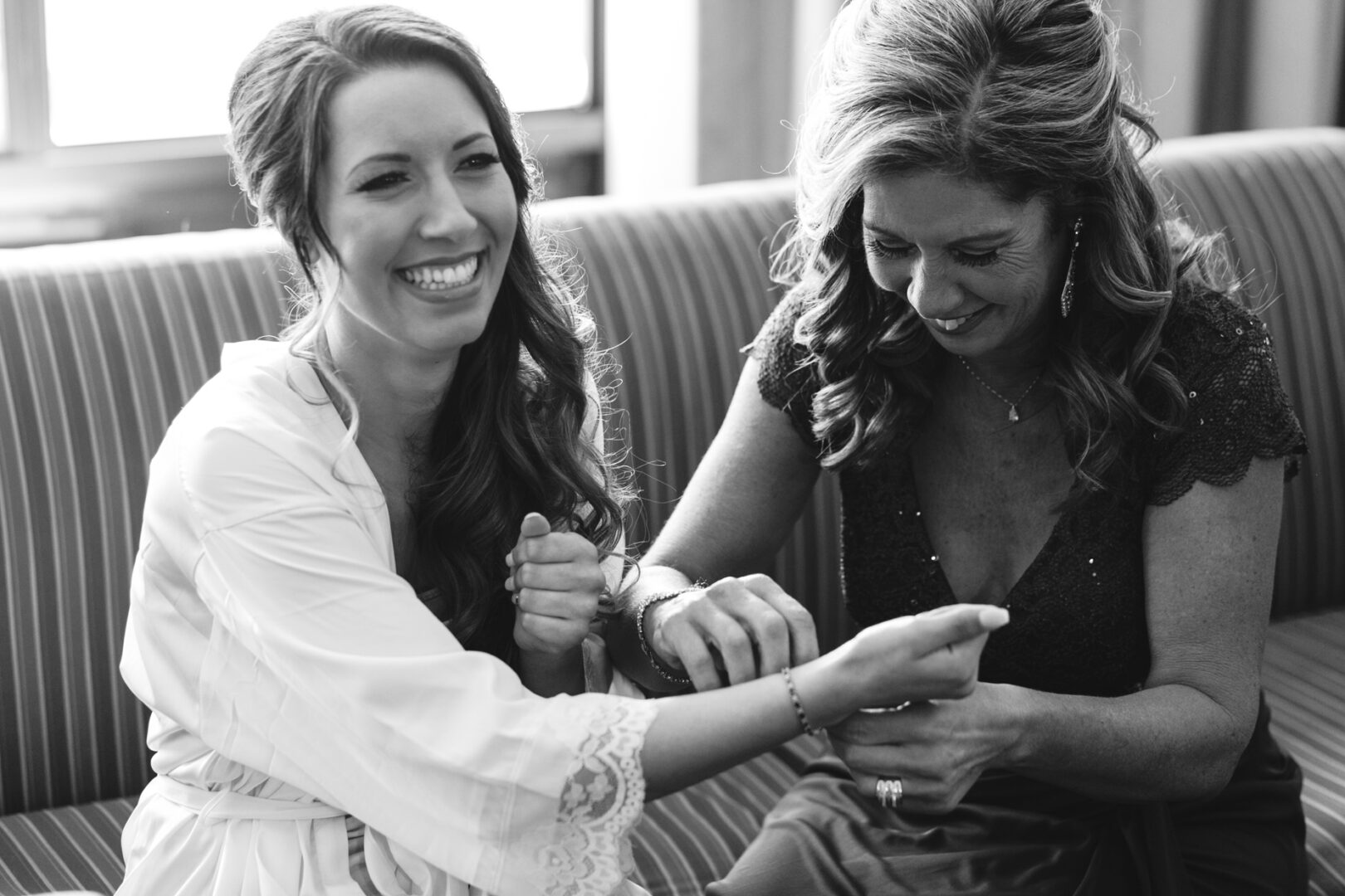 Two women sit on a striped couch, smiling and laughing like they just shared a dance at the Windsor Ballroom wedding. One adjusts the other's bracelet with care.