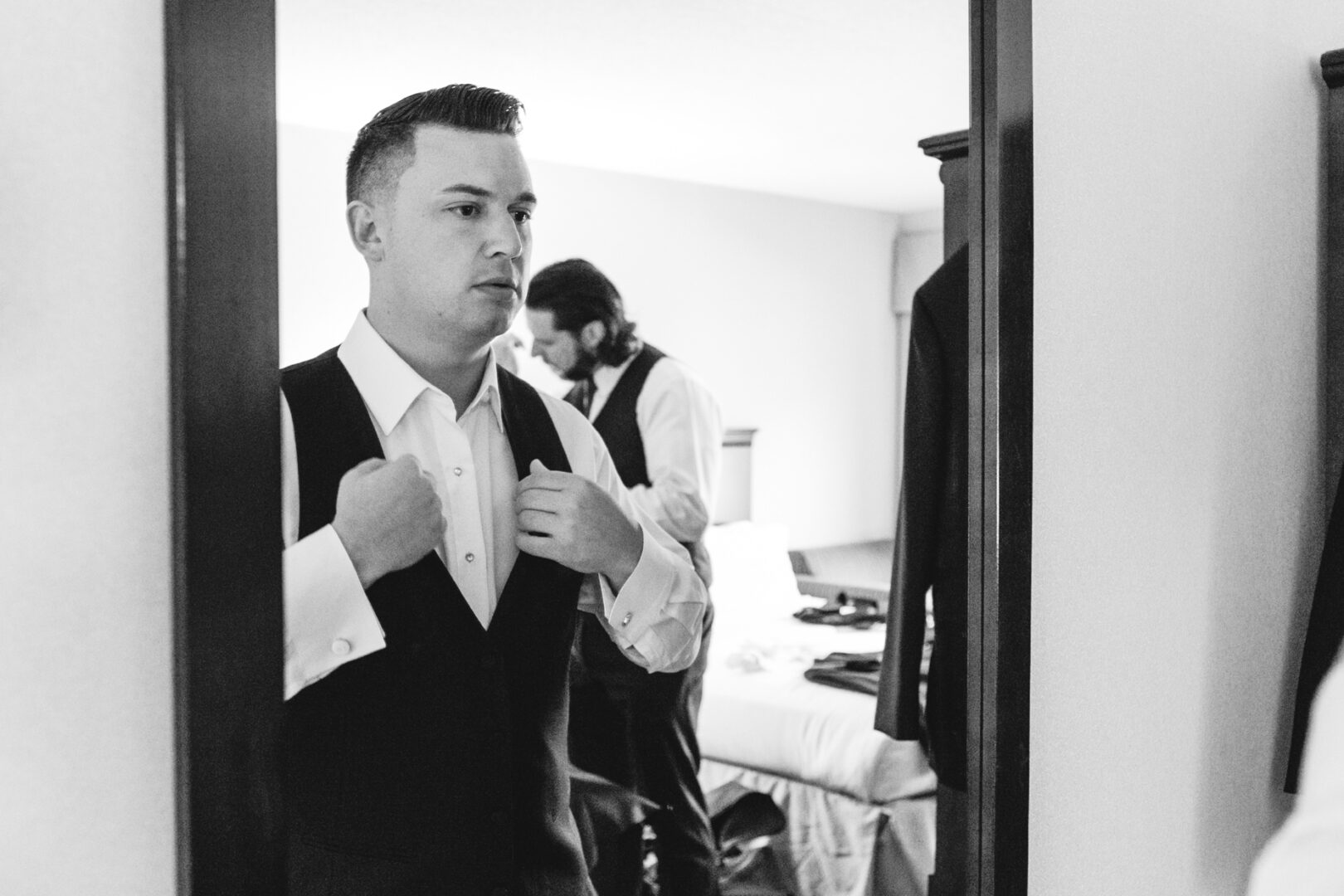 In the elegant Windsor Ballroom, a man adjusts his vest in front of a mirror while another stands nearby, both impeccably dressed for the wedding.