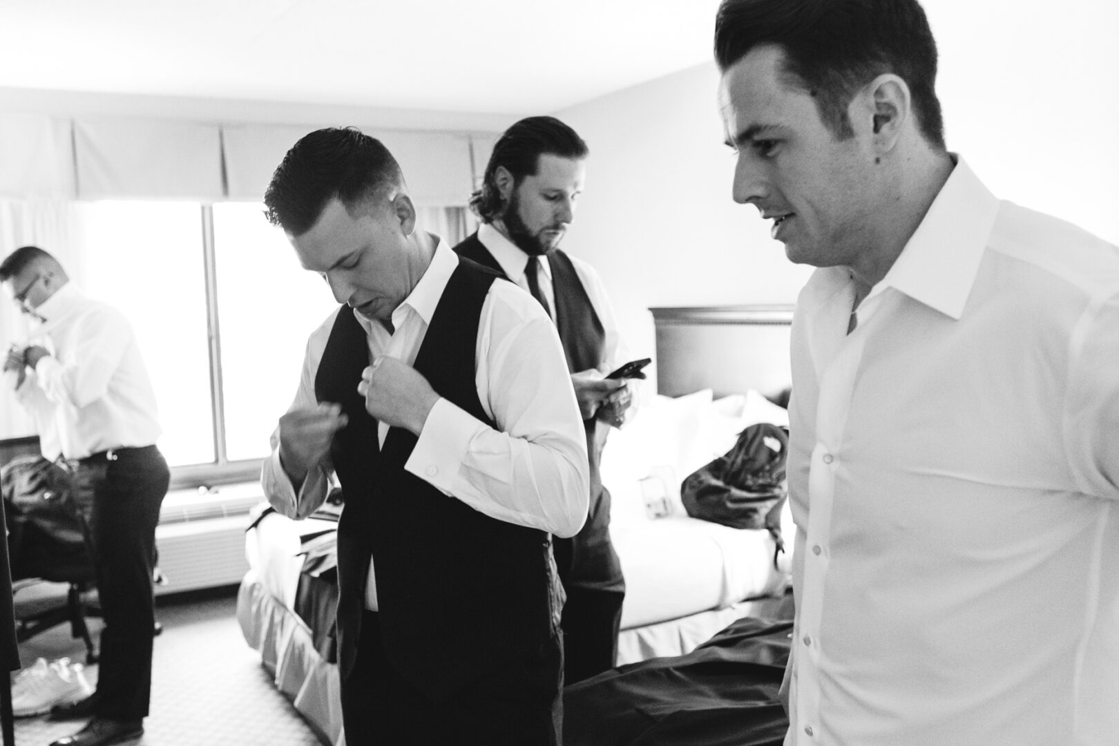 In the elegance of the Windsor Ballroom, four men prepare with precision—one adjusts his vest while another checks his phone, all in anticipation of the wedding's grandeur.