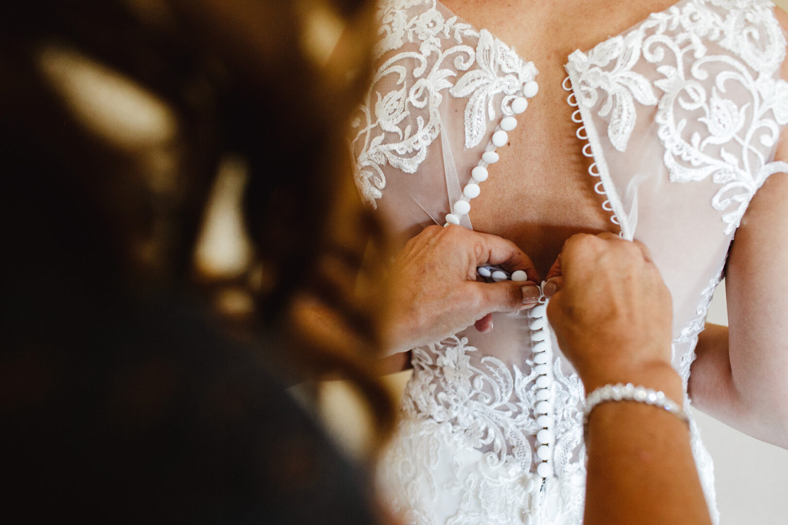 In the elegant setting of a Windsor Ballroom wedding, a person carefully fastens buttons on the back of a delicate lace wedding dress.