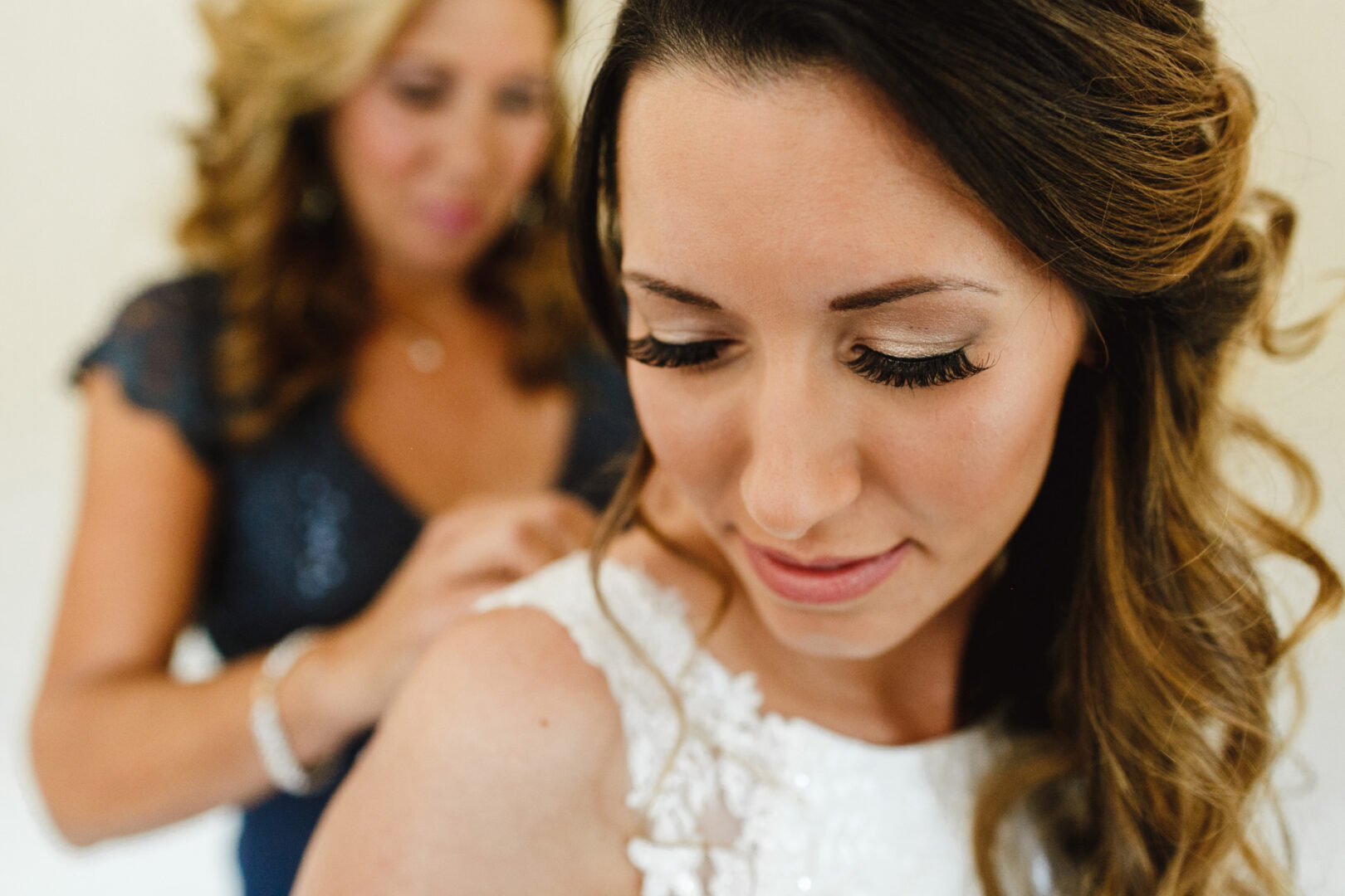 A woman in a white dress with her eyes closed is being helped by another woman in the background, preparing for her Windsor Ballroom wedding.