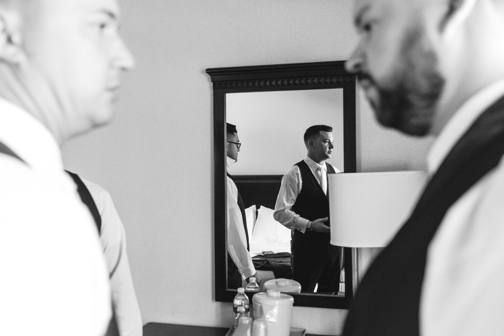 In a black-and-white scene, a man in a suit is reflected in a mirror, flanked by two others. They stand in what appears to be the Windsor Ballroom, where an elegant lamp casts soft shadows over the table adorned with bottles.