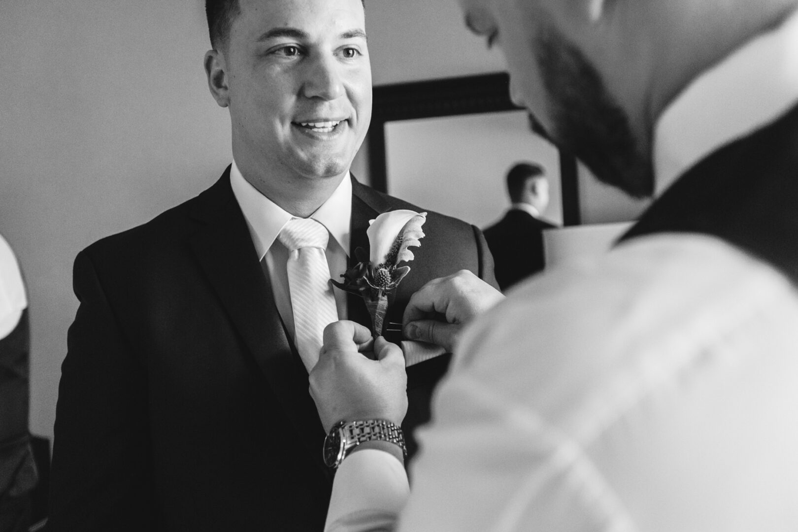 In a room preparing for a Windsor Ballroom wedding, a man carefully adjusts the boutonniere on another man's suit jacket, both dressed impeccably for the formal occasion.