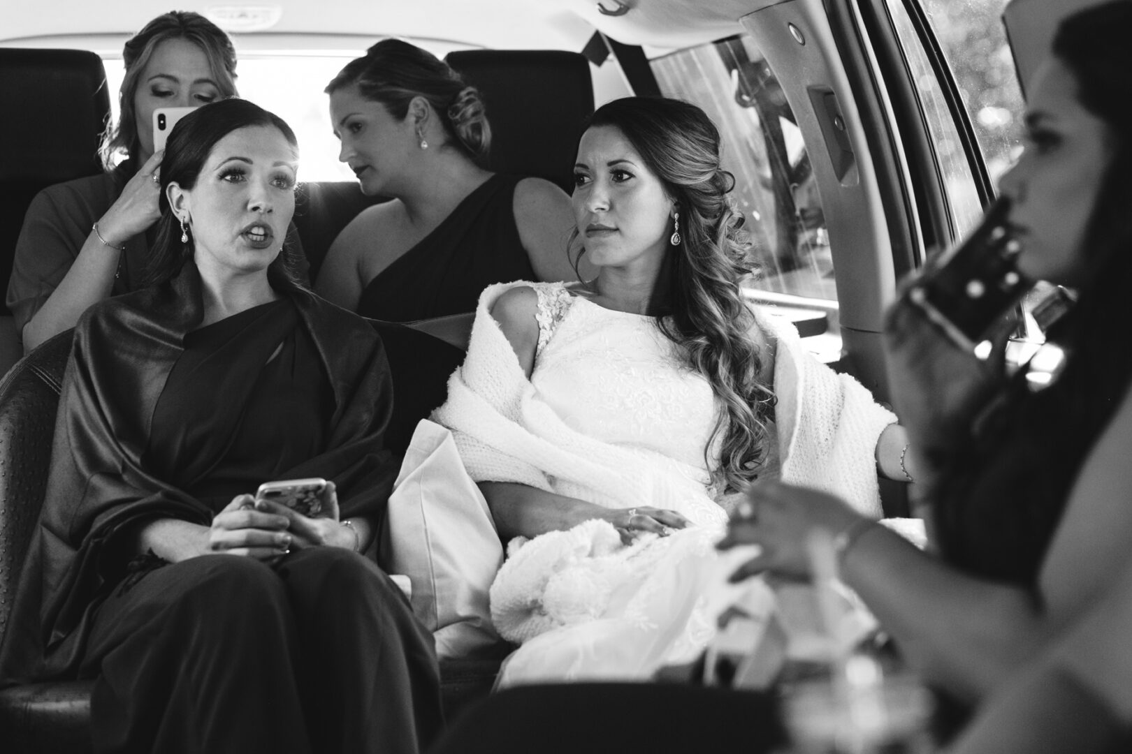 Inside the vehicle, a bride and her bridesmaids, draped in elegant formal attire, savor a serene moment before the Windsor Ballroom wedding. The bride, wrapped in a delicate shawl, gazes forward as soft daylight filters through while a bridesmaid animatedly converses beside her.