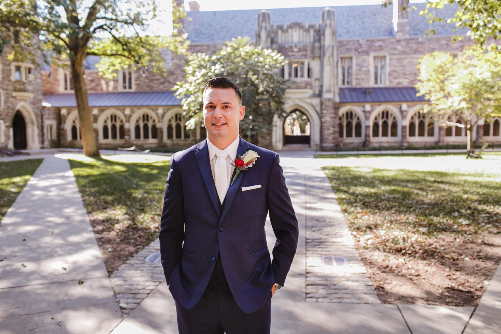 Man in a navy suit with a boutonniere stands outdoors on a pathway in front of a historic building, capturing the elegance reminiscent of a Windsor Ballroom wedding.