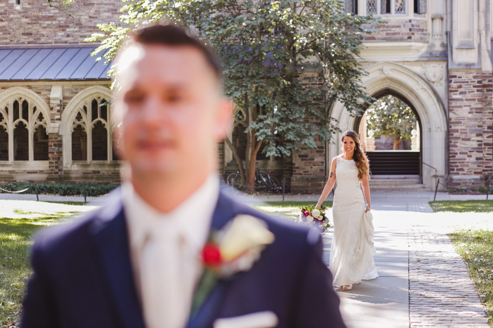 A bride walks towards a groom in a suit, standing outside the historic Windsor Ballroom, surrounded by archways and lush greenery.