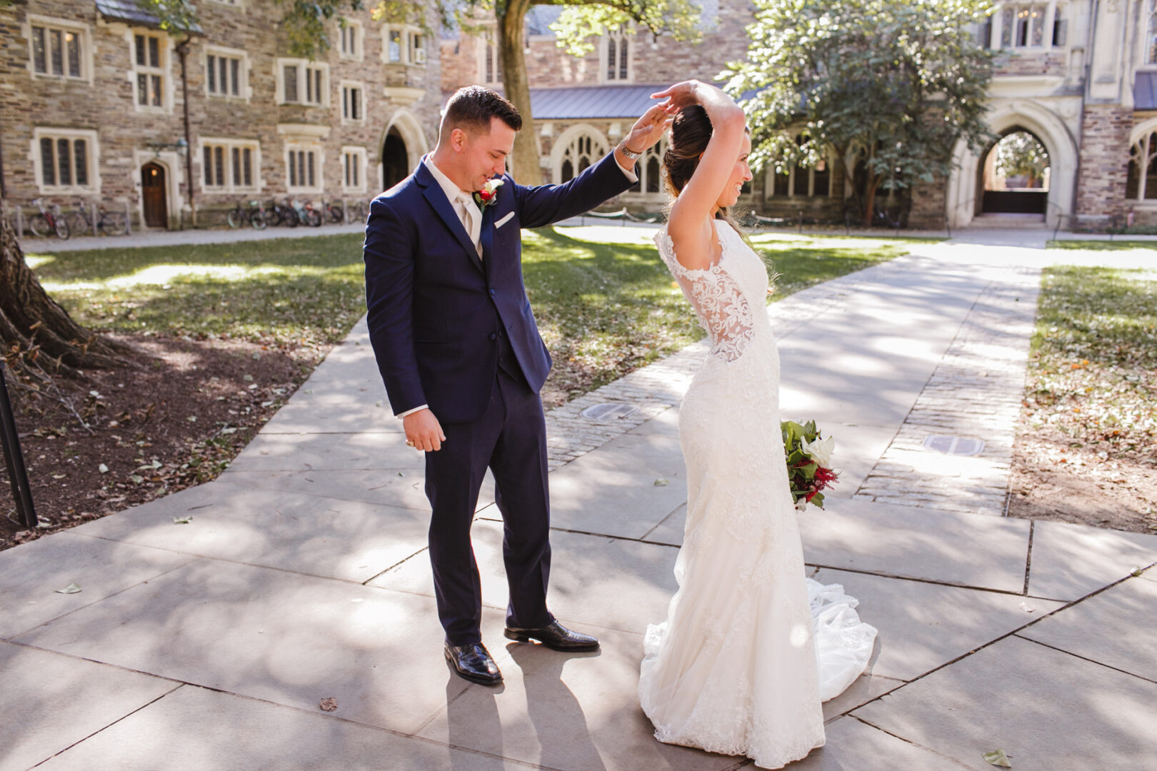 In the serene courtyard with stone buildings and a graceful tree, a groom in a dark suit twirls his bride, dressed in white and holding a bouquet. Their dance echoes with the elegance of a Windsor Ballroom wedding.