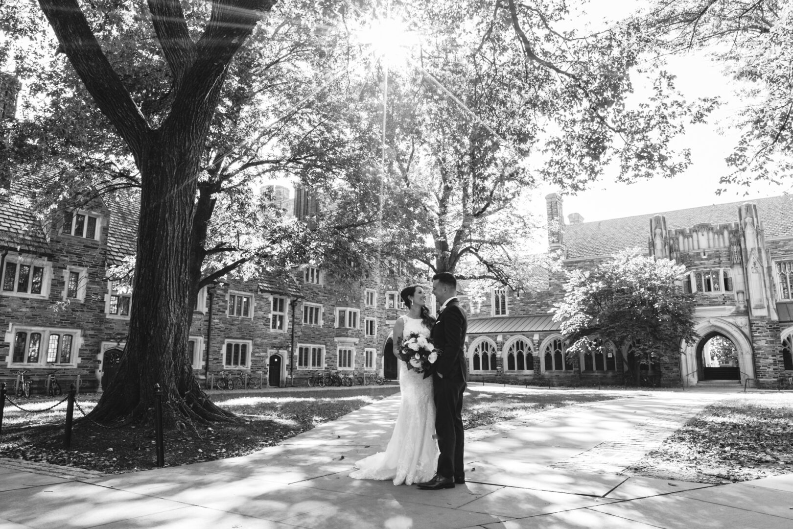 A couple stands together in wedding attire under large trees in a sunlit courtyard, reminiscent of a Windsor Ballroom Wedding, with historic buildings elegantly framing the backdrop.