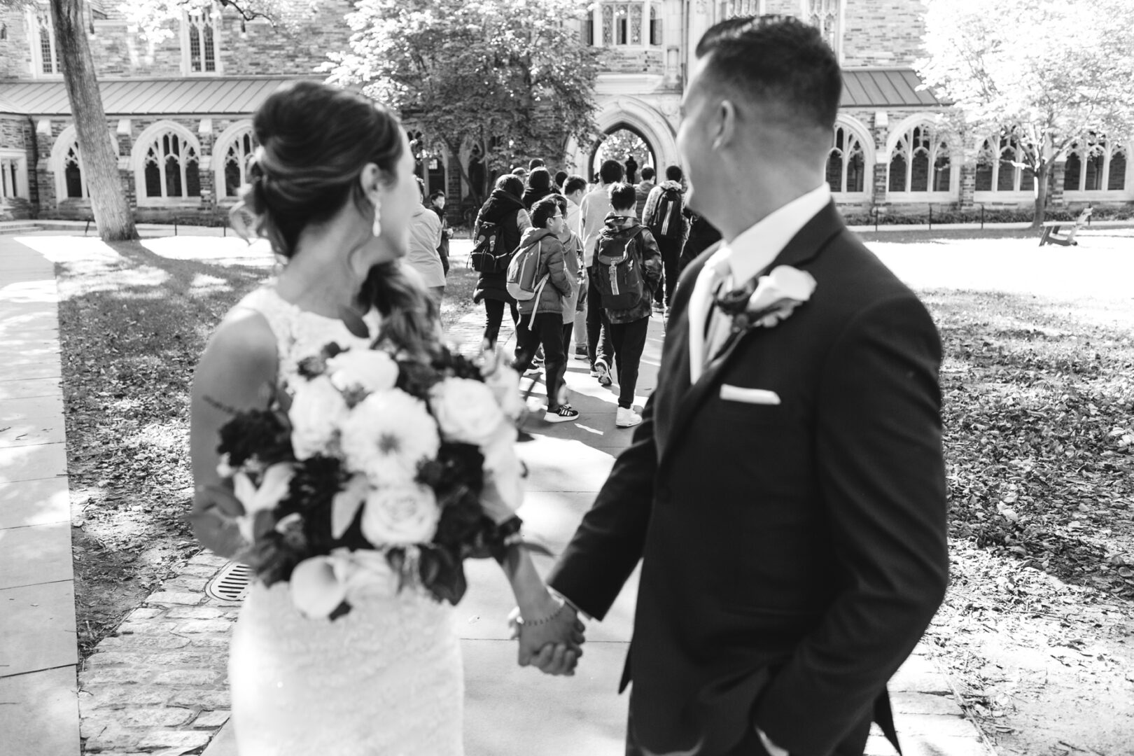 A bride and groom in wedding attire hold hands, gazing toward a group approaching the Windsor Ballroom, where their wedding celebration awaits.