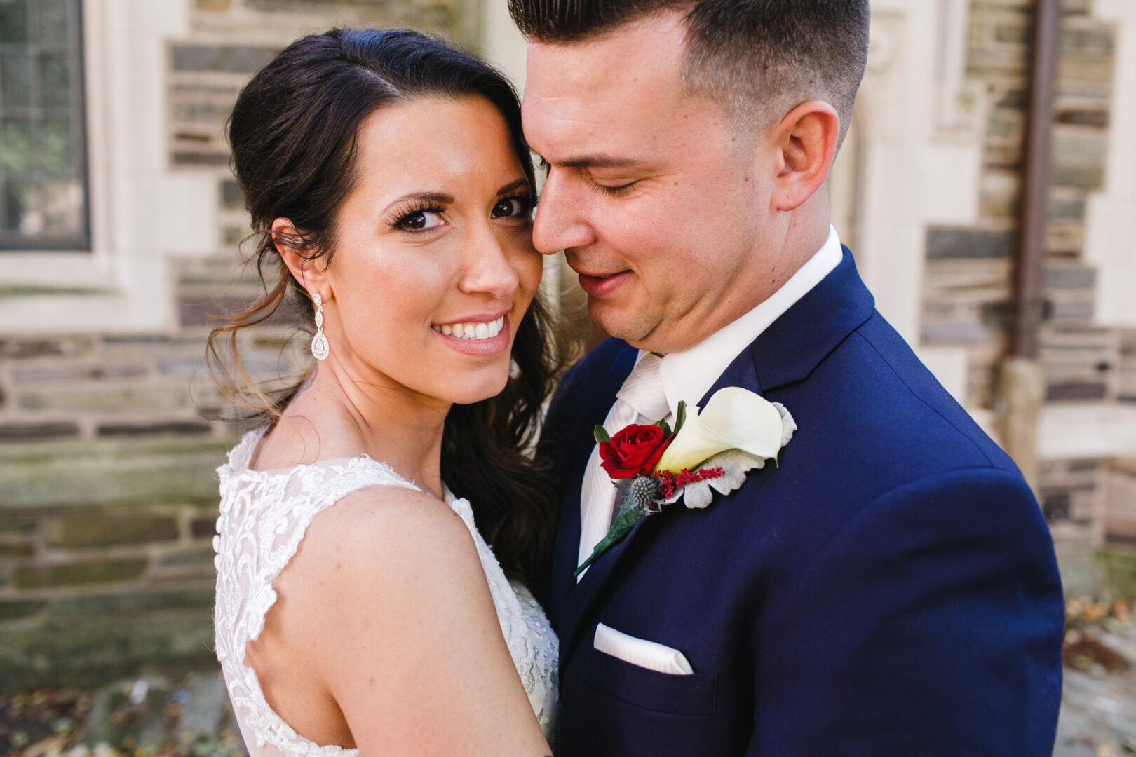 A couple embraces outdoors, with the woman smiling at the camera and the man looking at her lovingly. She wears a lace dress while he sports a navy suit adorned with a red and white boutonniere, capturing the essence of their enchanting Windsor Ballroom Wedding.