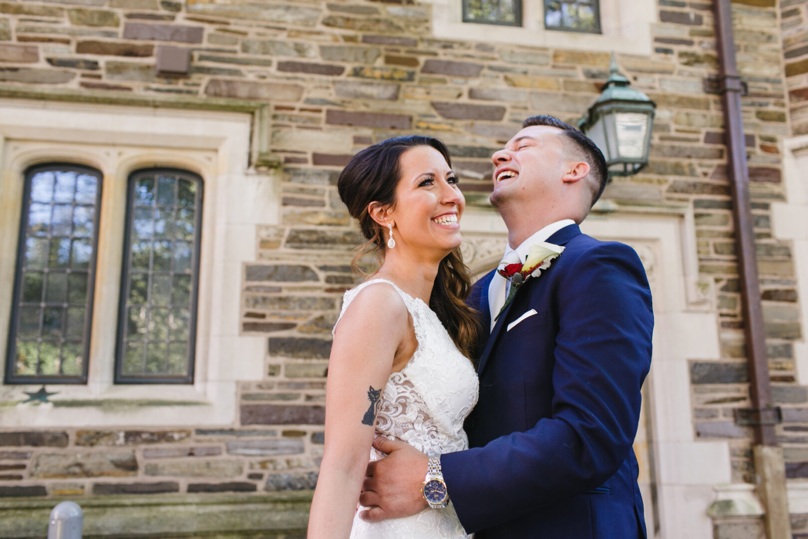 A couple in formal attire, caught in a moment of laughter, stands before the grand stone facade of the Windsor Ballroom, where large windows and a classic lantern set the perfect backdrop for their wedding day.