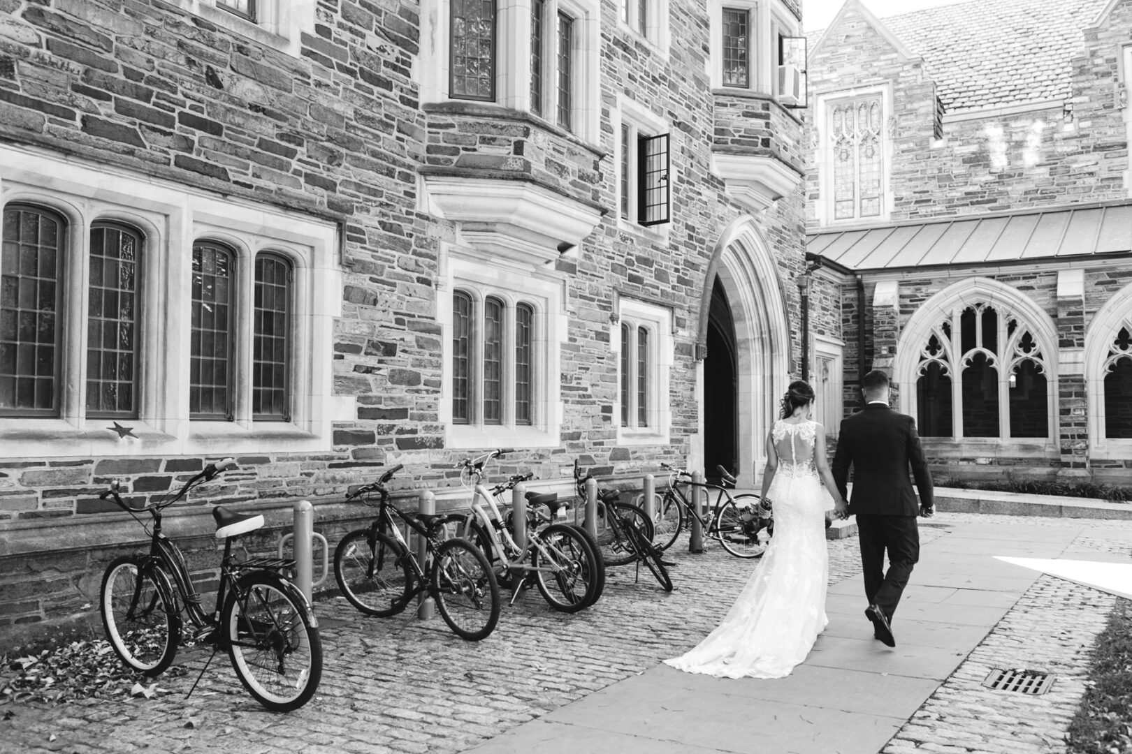 A couple strolls hand in hand along a cobblestone path, weaving between bikes as they approach the arched entrance of the brick building, dreaming of their Windsor Ballroom wedding.