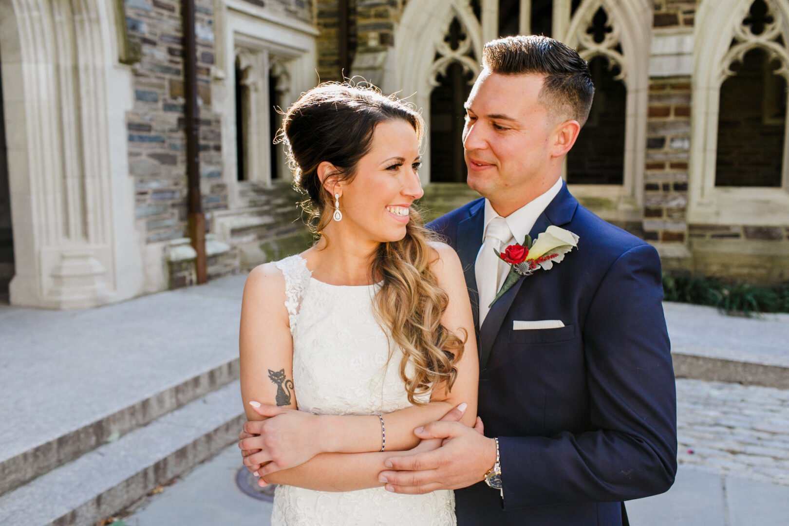 A couple embraces outside a stone building with gothic-style arches, reminiscent of a Windsor Ballroom Wedding. The woman is in a white dress, and the man, adorned with a red rose boutonniere on his suit, complements the elegance of the moment.