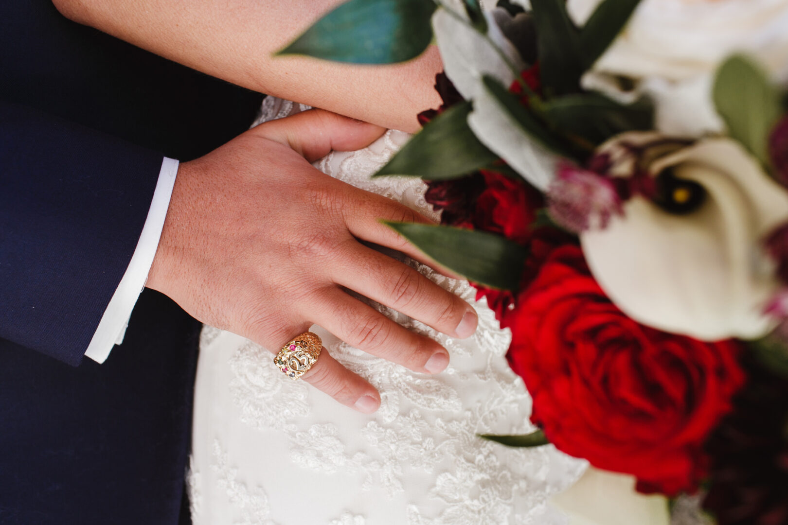 A hand with a gold ring rests on a woman's lace dress, holding a bouquet with red and white flowers, as they prepare for their Windsor Ballroom wedding.