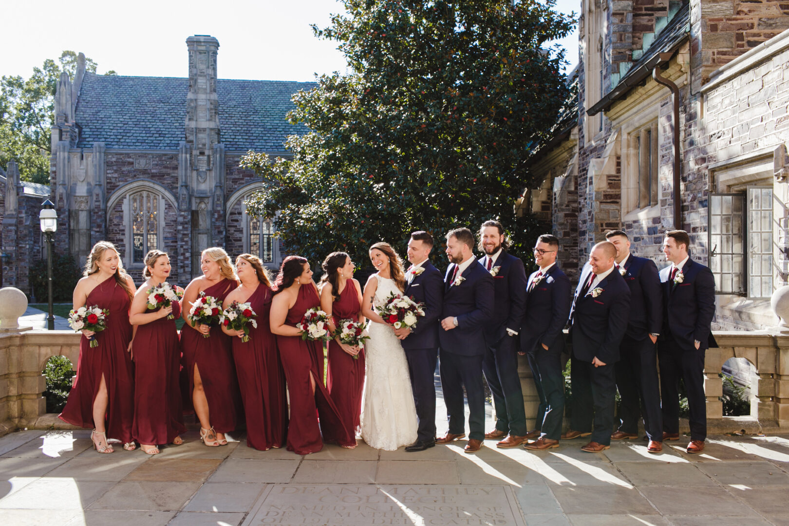 A bride and groom pose with their bridesmaids and groomsmen in formal attire outside a stone building on a sunny day, reminiscent of an elegant Windsor Ballroom wedding. Bridesmaids don maroon dresses, while the groomsmen are in dark suits.