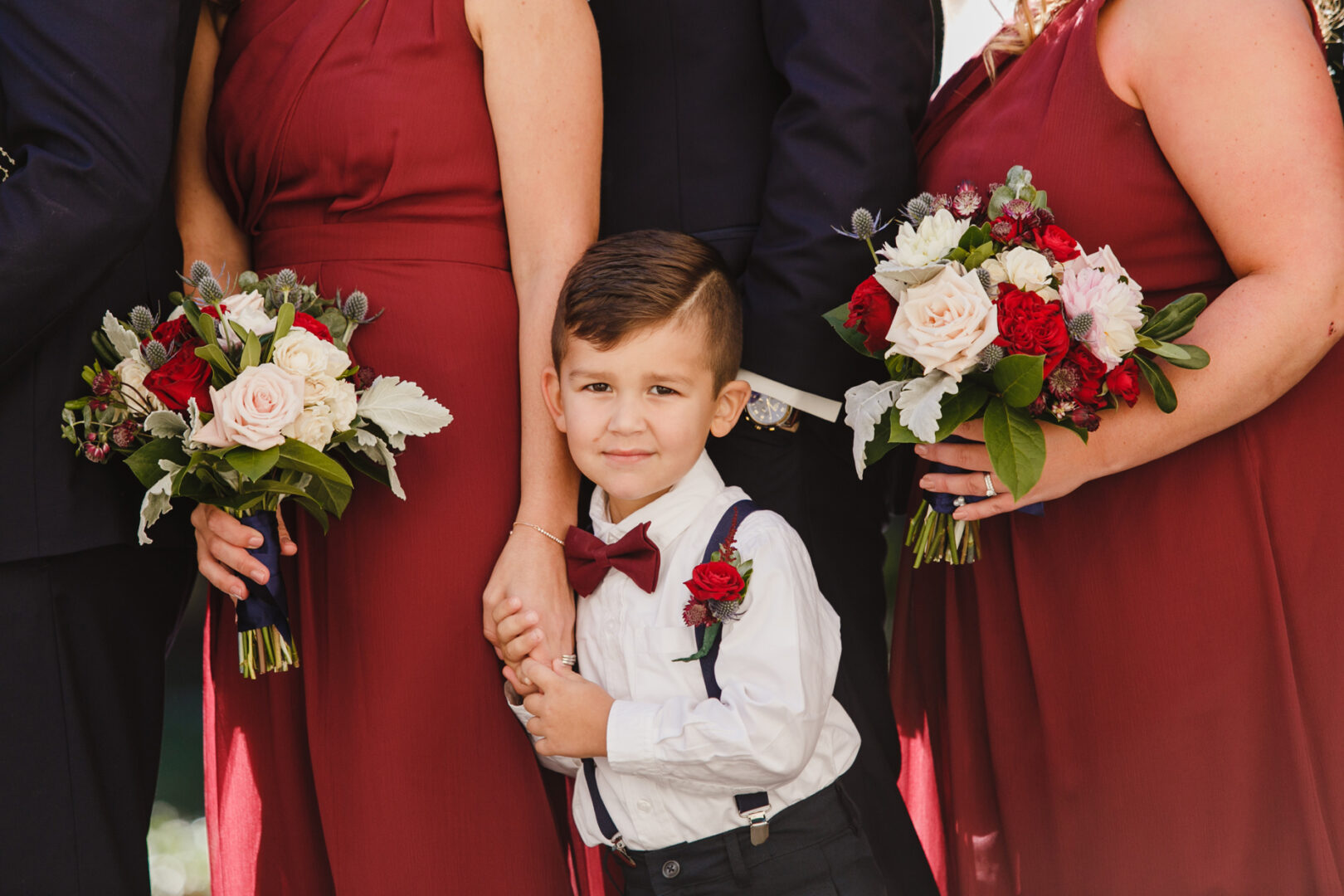 A young boy in a white shirt and red bow tie holds hands with adults in formal attire at a Windsor Ballroom wedding. They each hold bouquets of white and red flowers, adding a touch of elegance to the enchanting celebration.