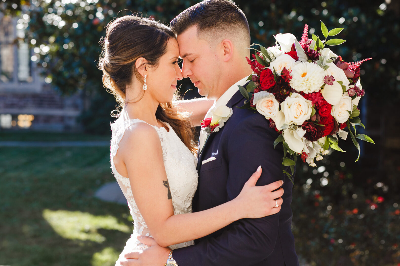 A bride and groom embrace outdoors, touching foreheads beneath the open sky, with a bouquet of red and white flowers—capturing the romance reminiscent of a Windsor Ballroom Wedding.