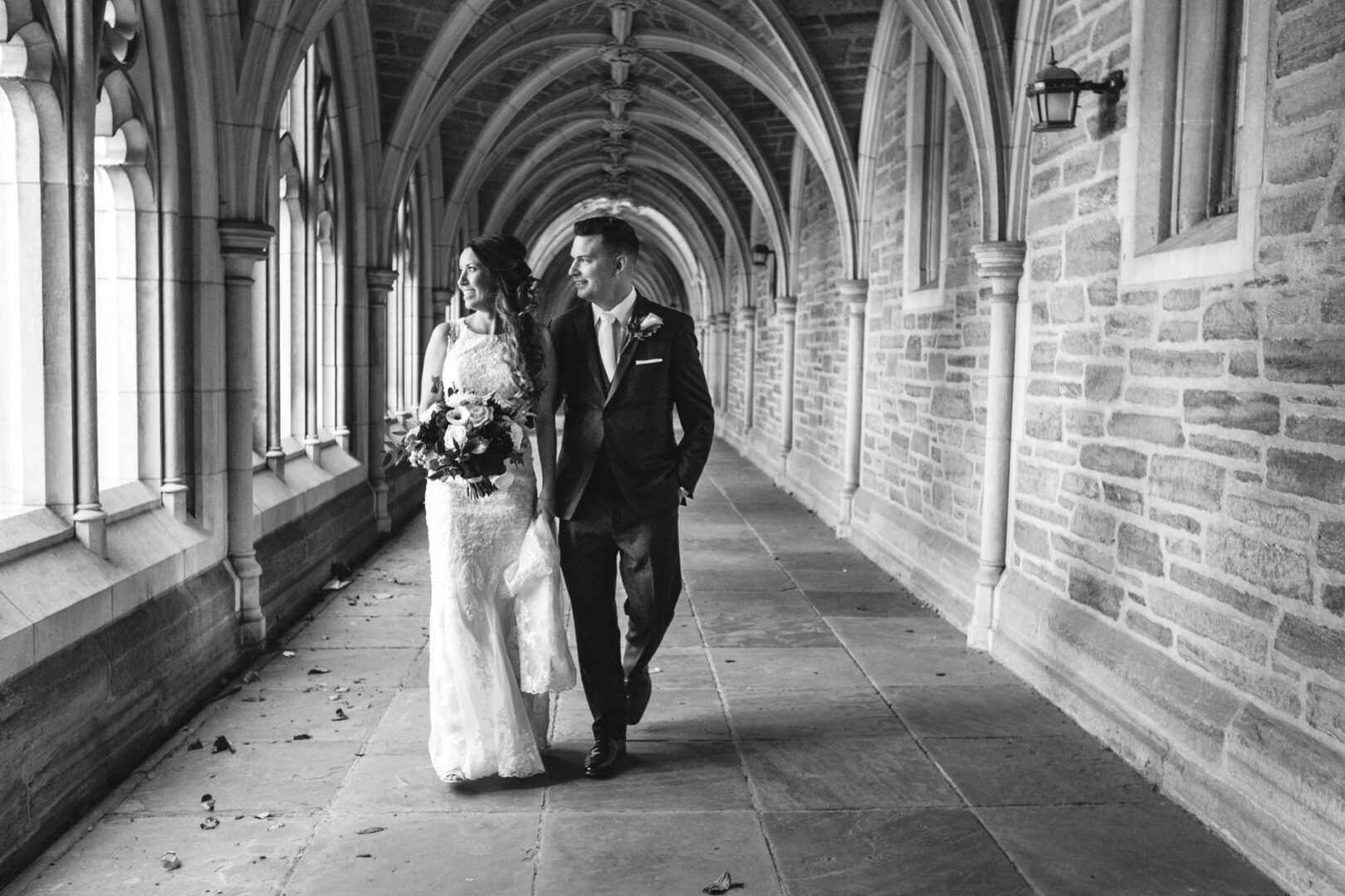 A couple in formal attire strolls through an arched stone corridor, the woman clutching a bouquet, capturing the timeless elegance of a Windsor Ballroom wedding. The black and white scene highlights their grace and sophistication.