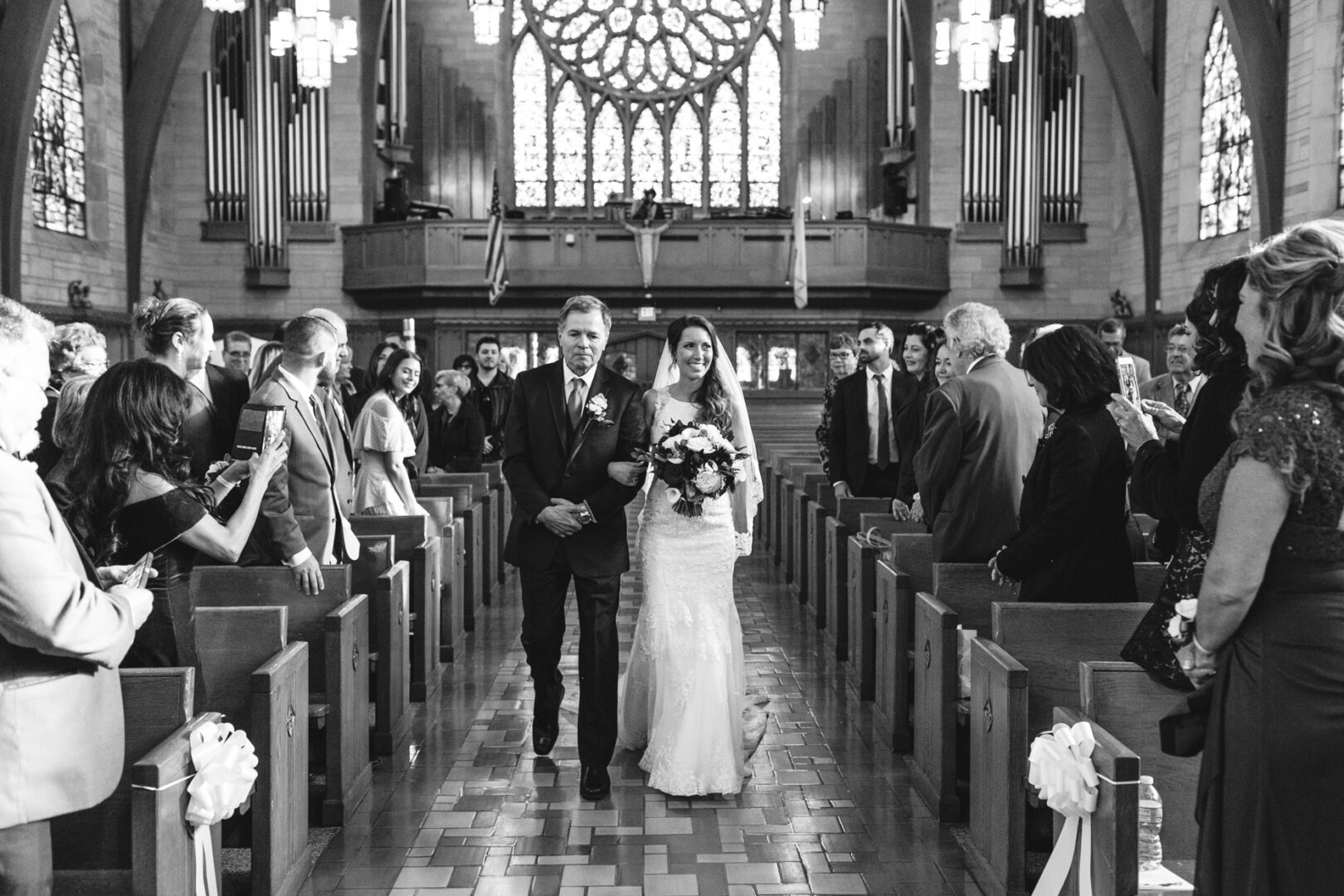 A bride, holding a bouquet, gracefully walks down the aisle of the Windsor Ballroom with an older man. Guests stand on either side in awe, capturing the moment with their phones.