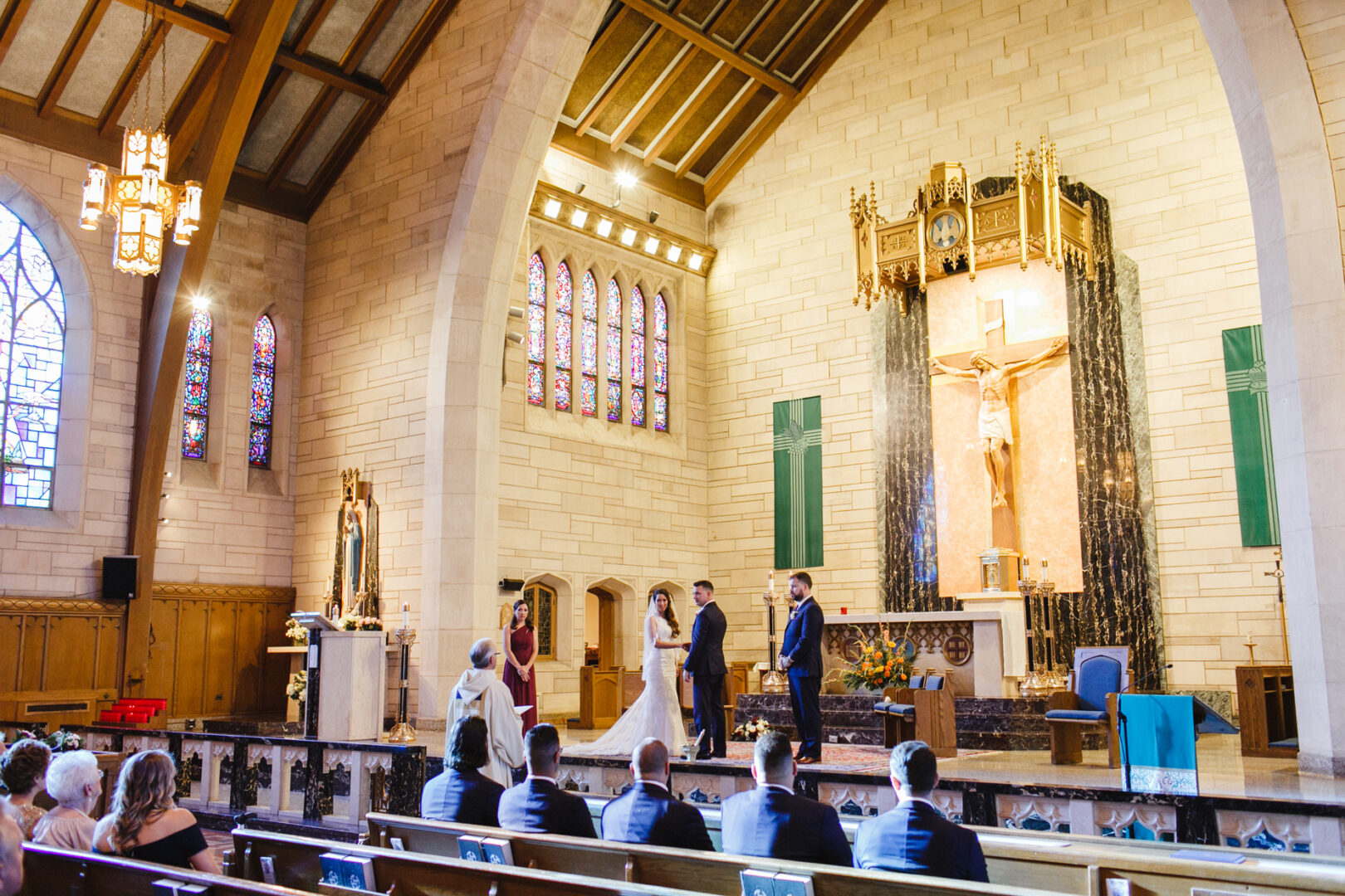 A couple exchanges vows at the altar in a church, their eyes gleaming with love. The priest stands nearby, and guests are seated in the pews. The sacred air mingles with memories of their enchanting Windsor Ballroom wedding, as stained glass windows cast colorful reflections beside the crucifix.