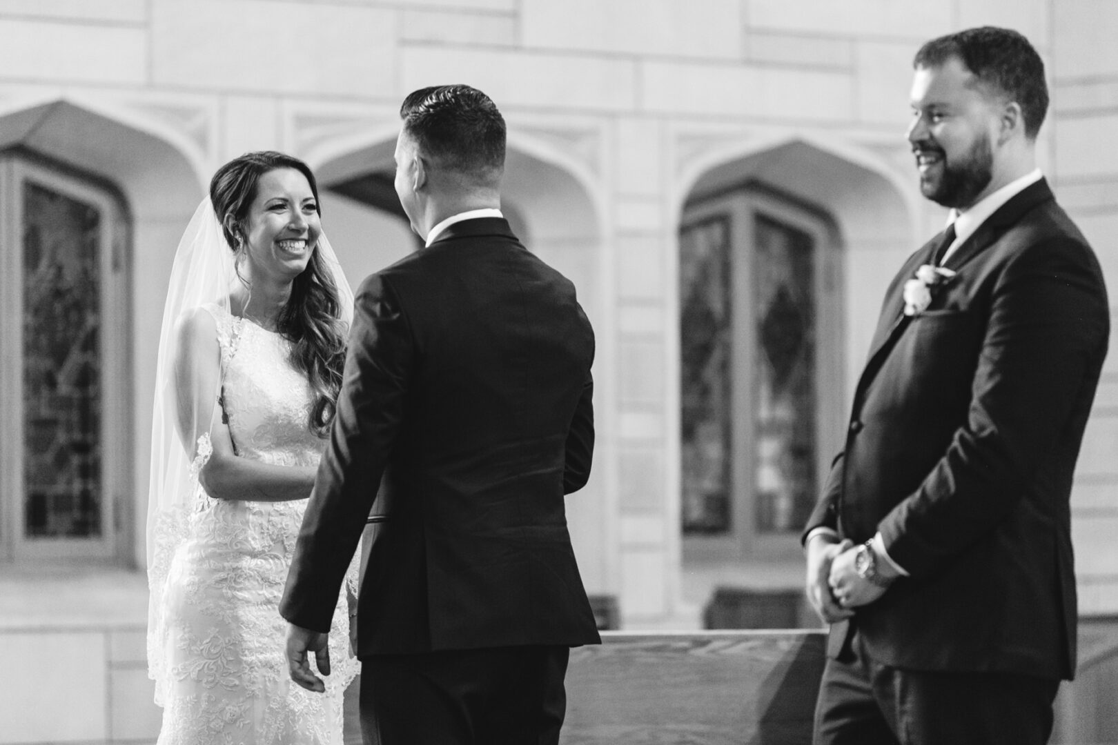 The bride smiles at the groom during their wedding ceremony in the church, while a groomsman stands nearby, capturing the elegance of a Windsor Ballroom wedding.