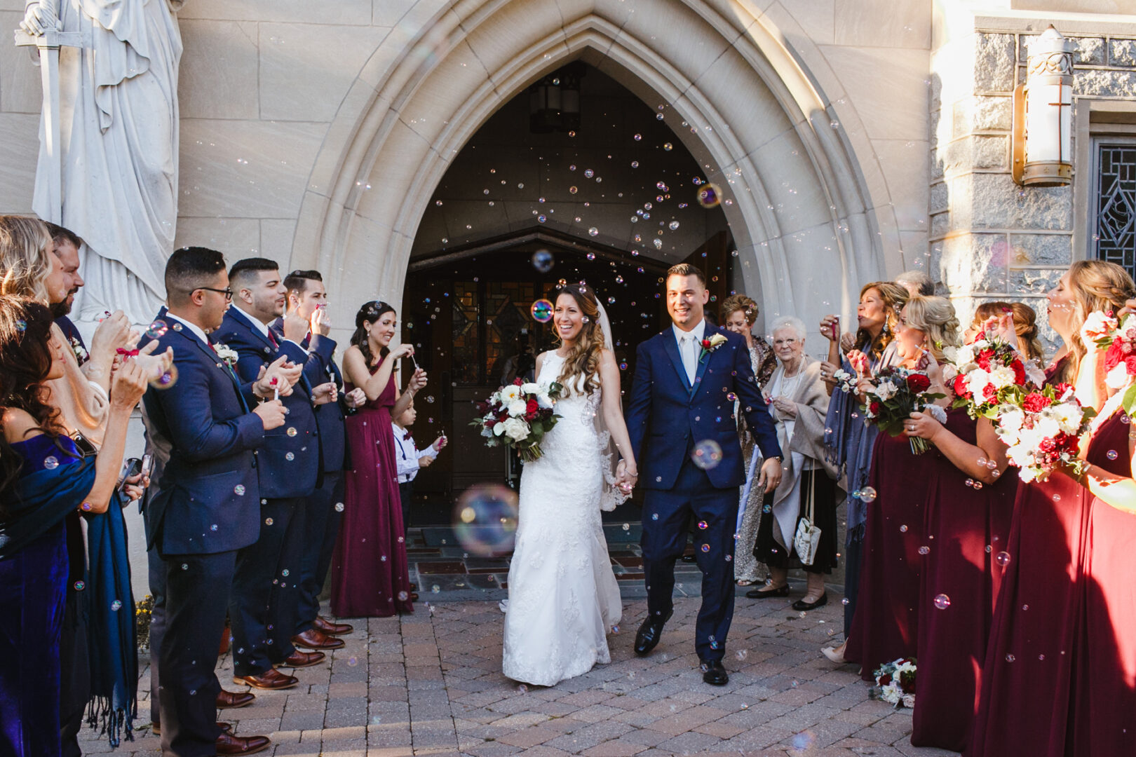 A bride and groom exit a church as guests blow bubbles and cheer, setting the festive tone for the Windsor Ballroom wedding reception. Bridesmaids in burgundy dresses and groomsmen in navy suits stand alongside, celebrating this beautiful union.