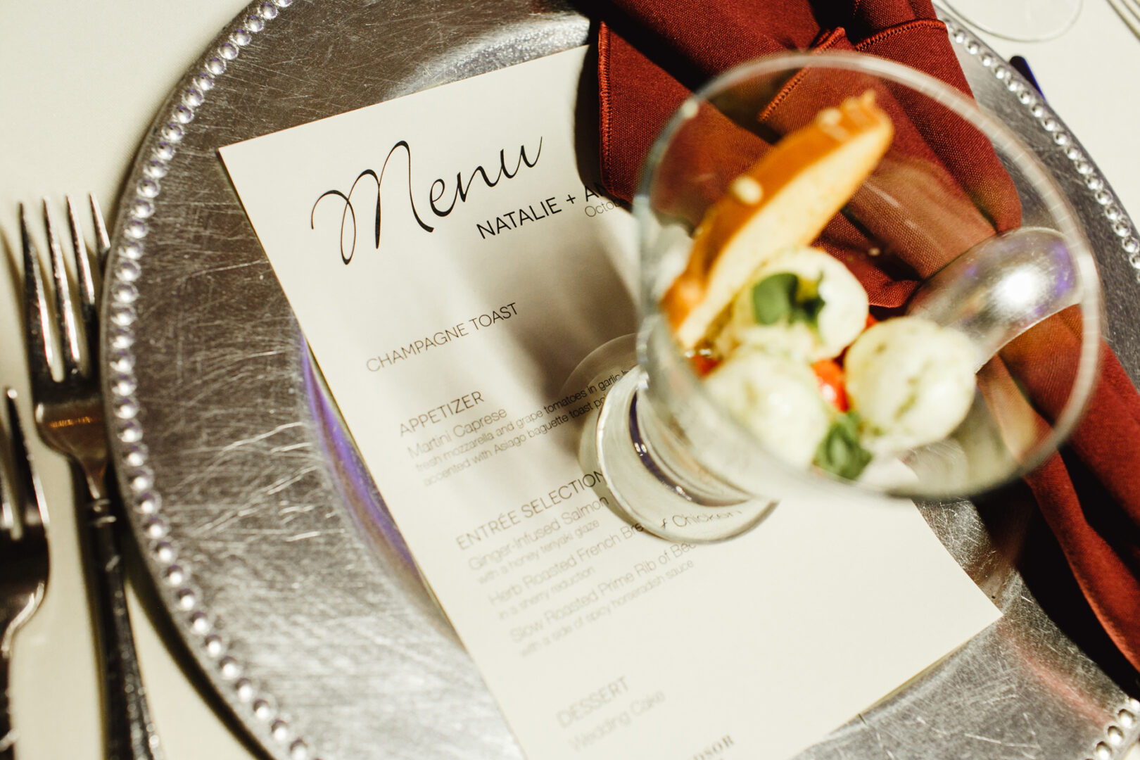 A table set for a Windsor Ballroom Wedding features a menu card elegantly placed on a silver plate. A glass filled with bread and mozzarella balls rests atop the menu, while a folded red napkin and cutlery are meticulously arranged beside it.