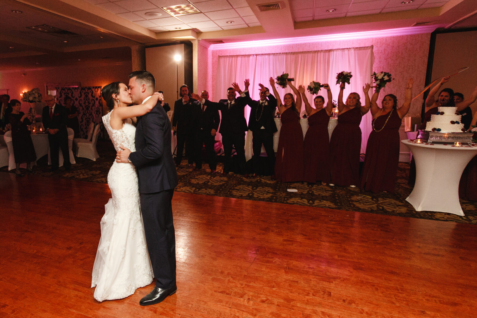 At a Windsor Ballroom Wedding, the bride and groom share their first dance as cheerful bridesmaids and groomsmen cheer them on in the background.