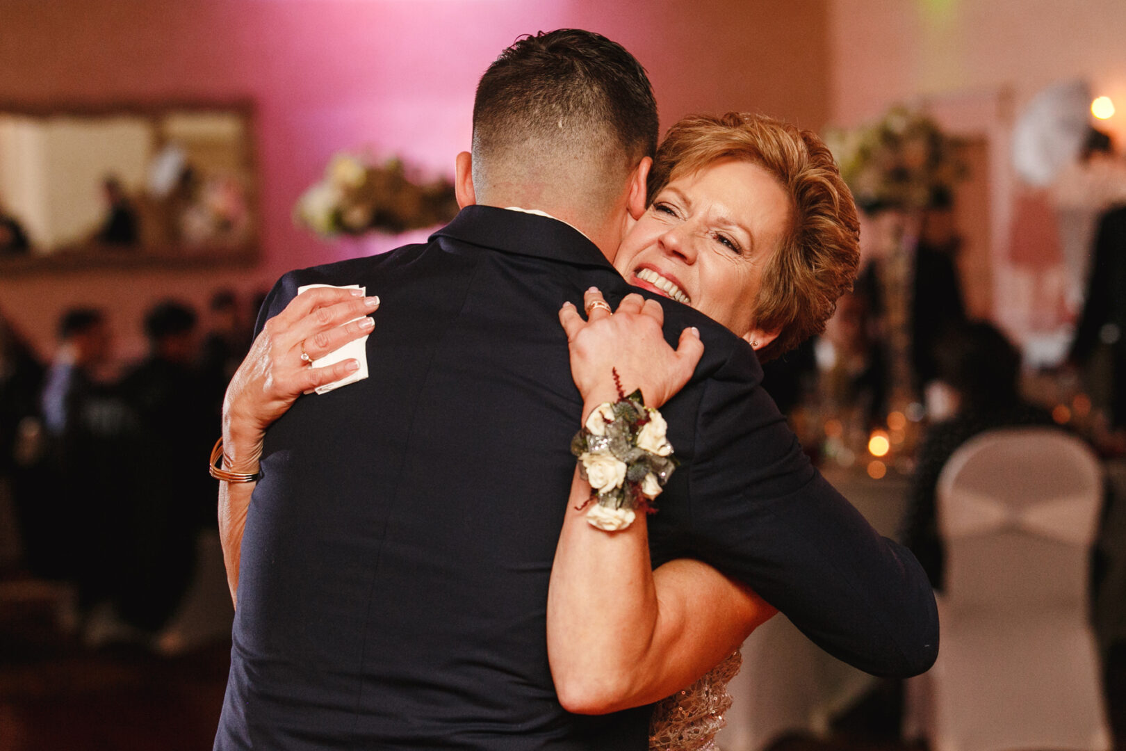 At the elegant Windsor Ballroom, a woman with a corsage hugs a man in a suit at the wedding, sharing a moment of genuine affection amidst the formal event.