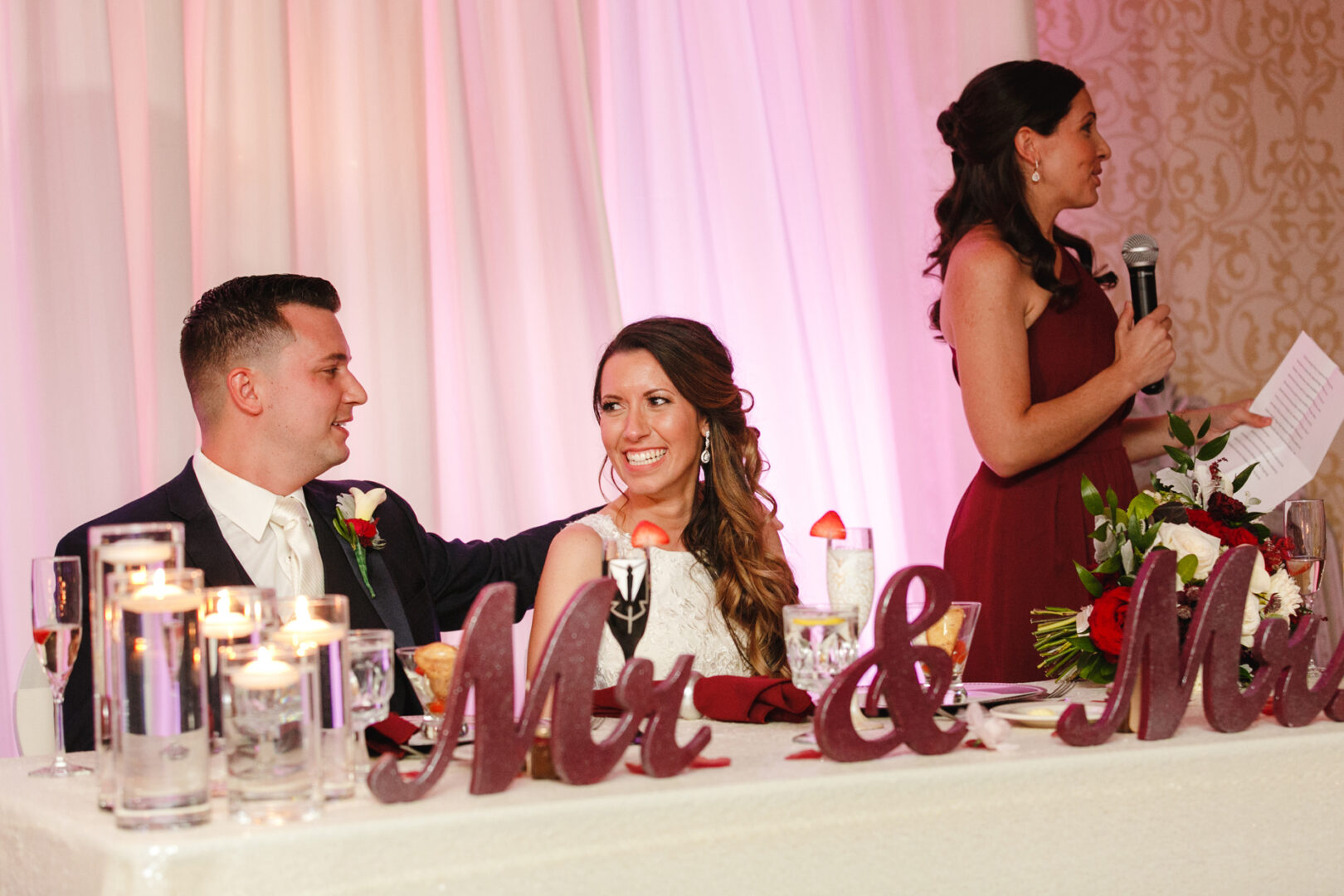 At the elegant Windsor Ballroom Wedding, the bride and groom sit at a reception table adorned with a "Mr. & Mrs." sign as a woman gracefully speaks into the microphone, adding warmth to the celebration.