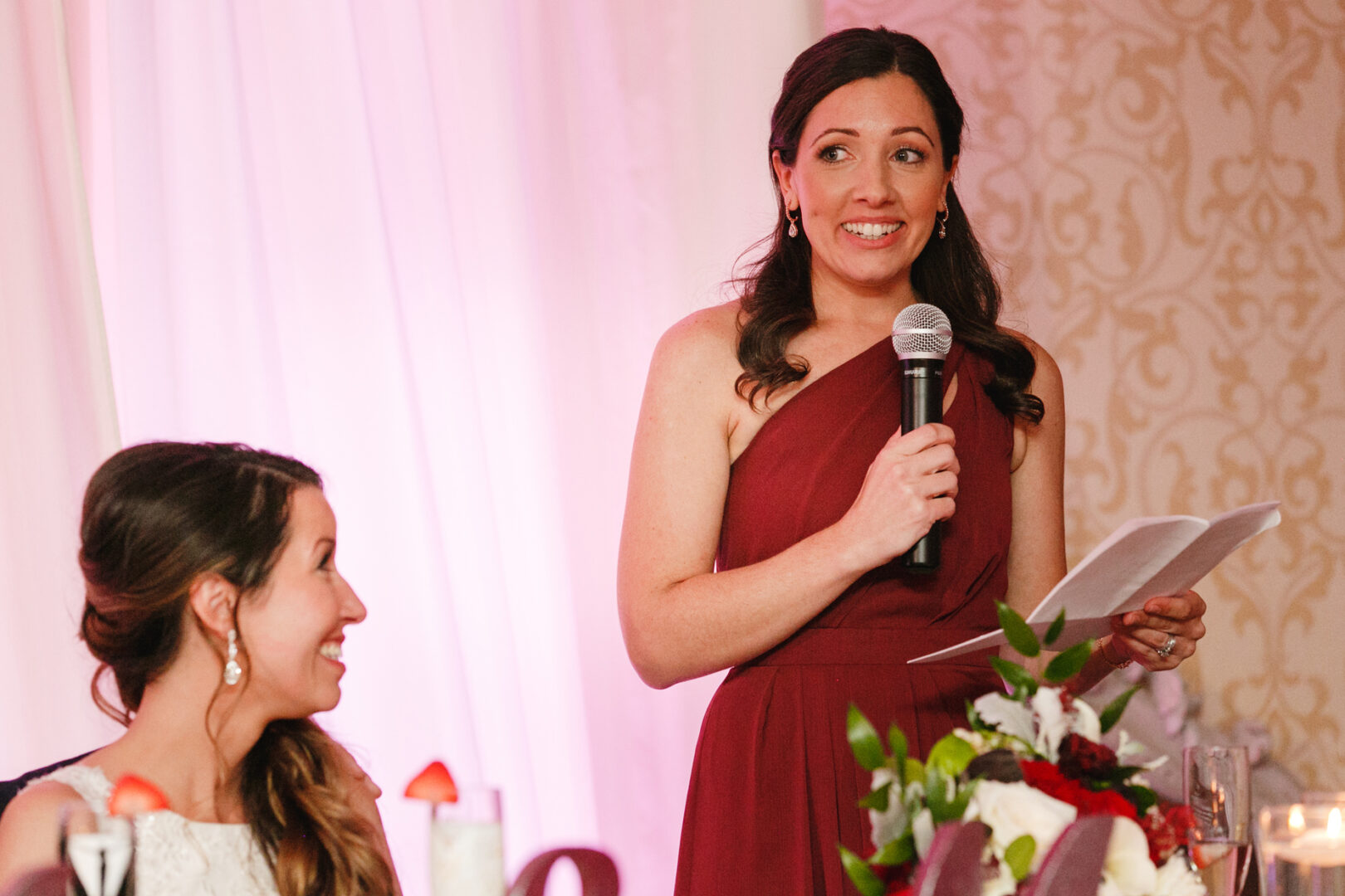 In the elegant Windsor Ballroom, a woman in a burgundy dress addresses the guests through a microphone, while another admires her amidst stunning floral decorations, crafting an unforgettable wedding atmosphere.