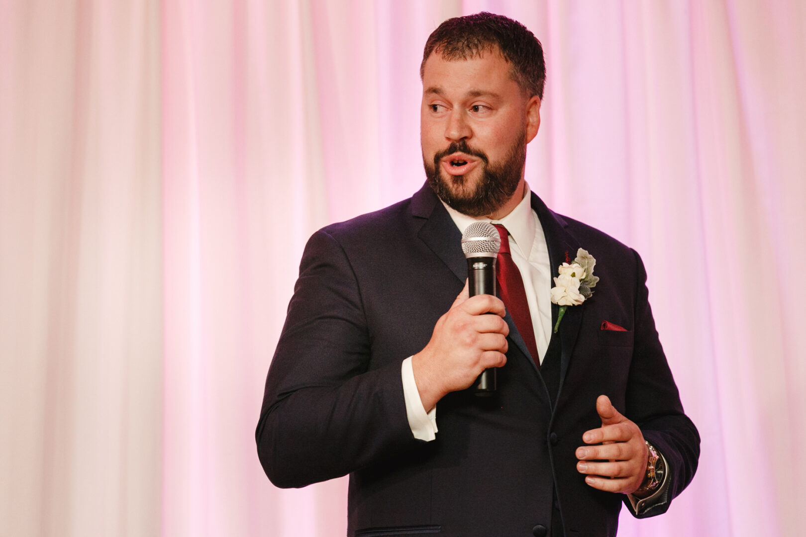 A man in a dark suit holds a microphone, poised to speak at what must be a delightful Windsor Ballroom wedding, with the pink and white draped backdrop adding an air of elegance.